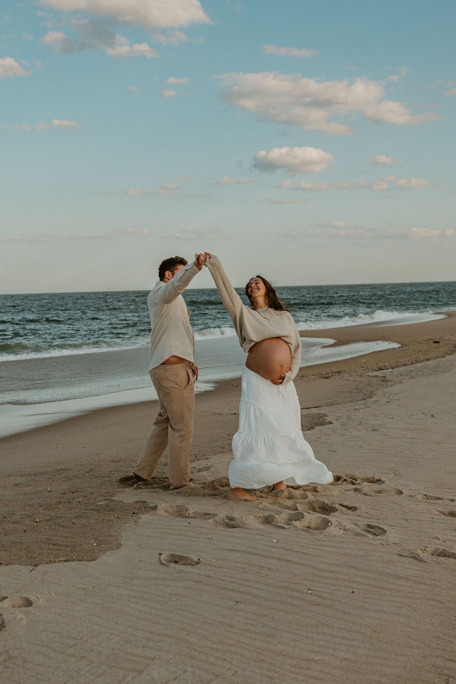 Couple dancing on beach at sunset, pregnant woman in white skirt holding hands with partner near ocean waves.