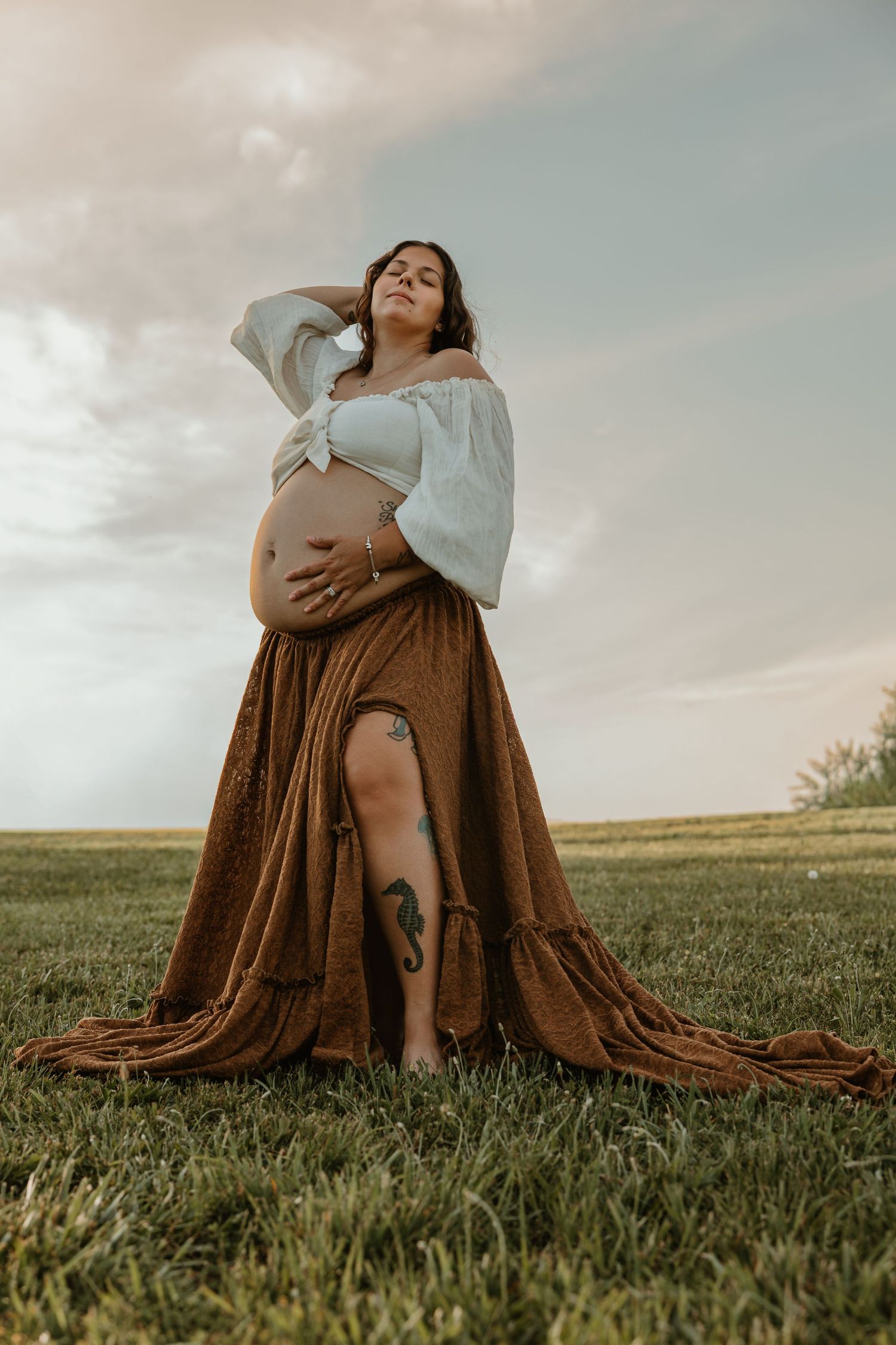 Pregnant woman in boho off-shoulder crop top and brown maxi skirt with leg tattoo poses outdoors on grass.