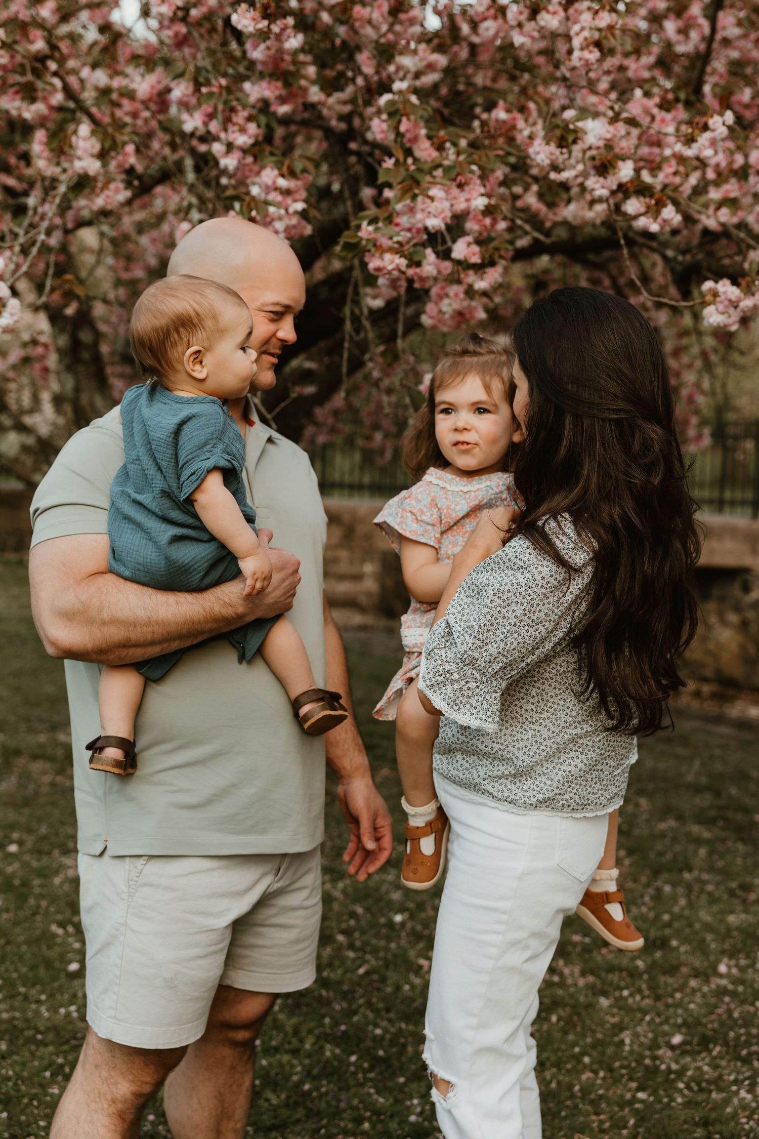 A happy family with two young children poses under pink cherry blossom trees in a spring outdoor portrait.