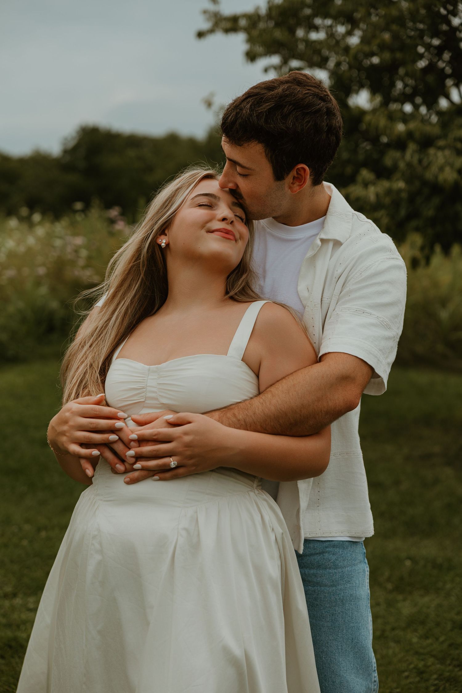A couple embraces outdoors, woman in white dress smiling as man kisses her head in a lush green garden setting.