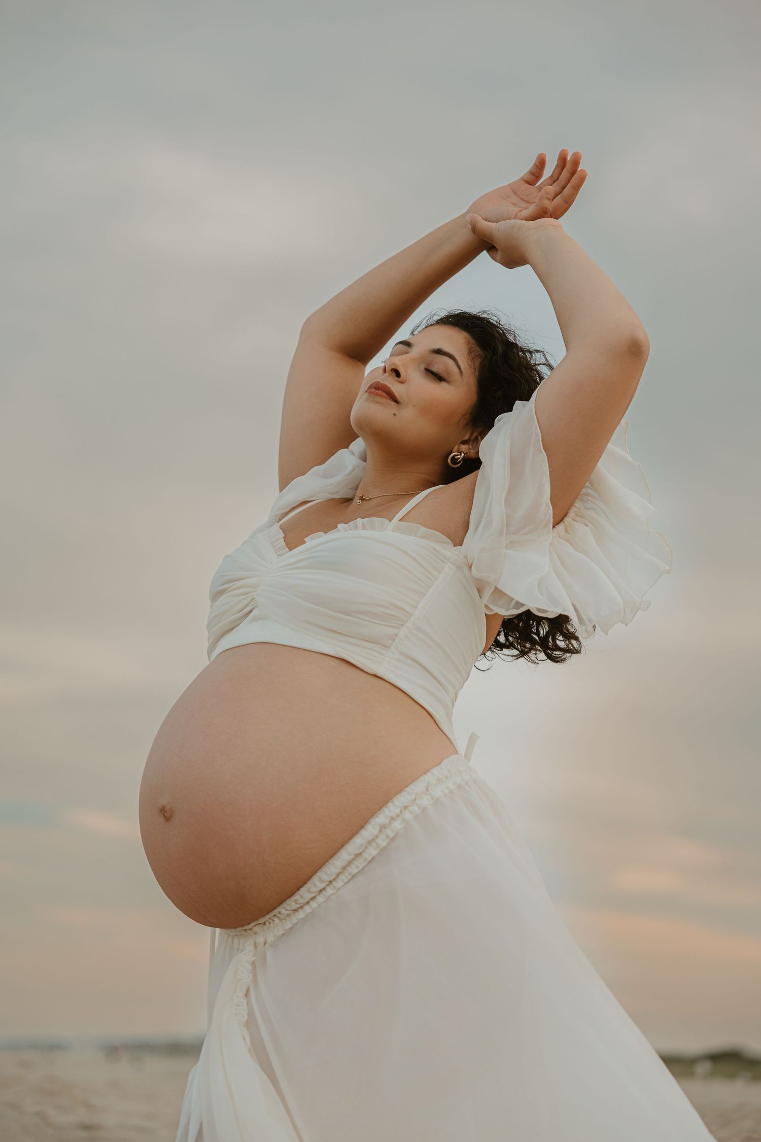 Pregnant woman in white crop top and skirt poses gracefully on beach at sunset with arms raised overhead.