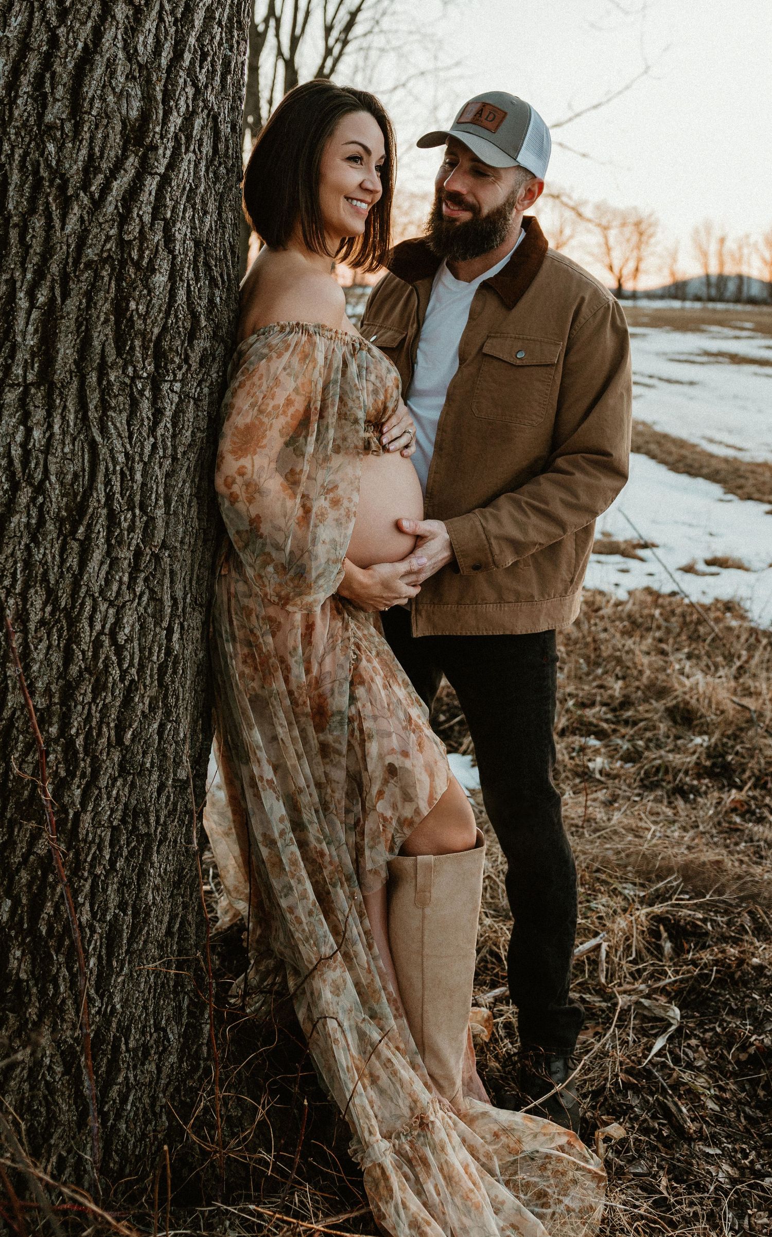 A romantic winter maternity photoshoot with a couple embracing near a tree in the snow at sunset.