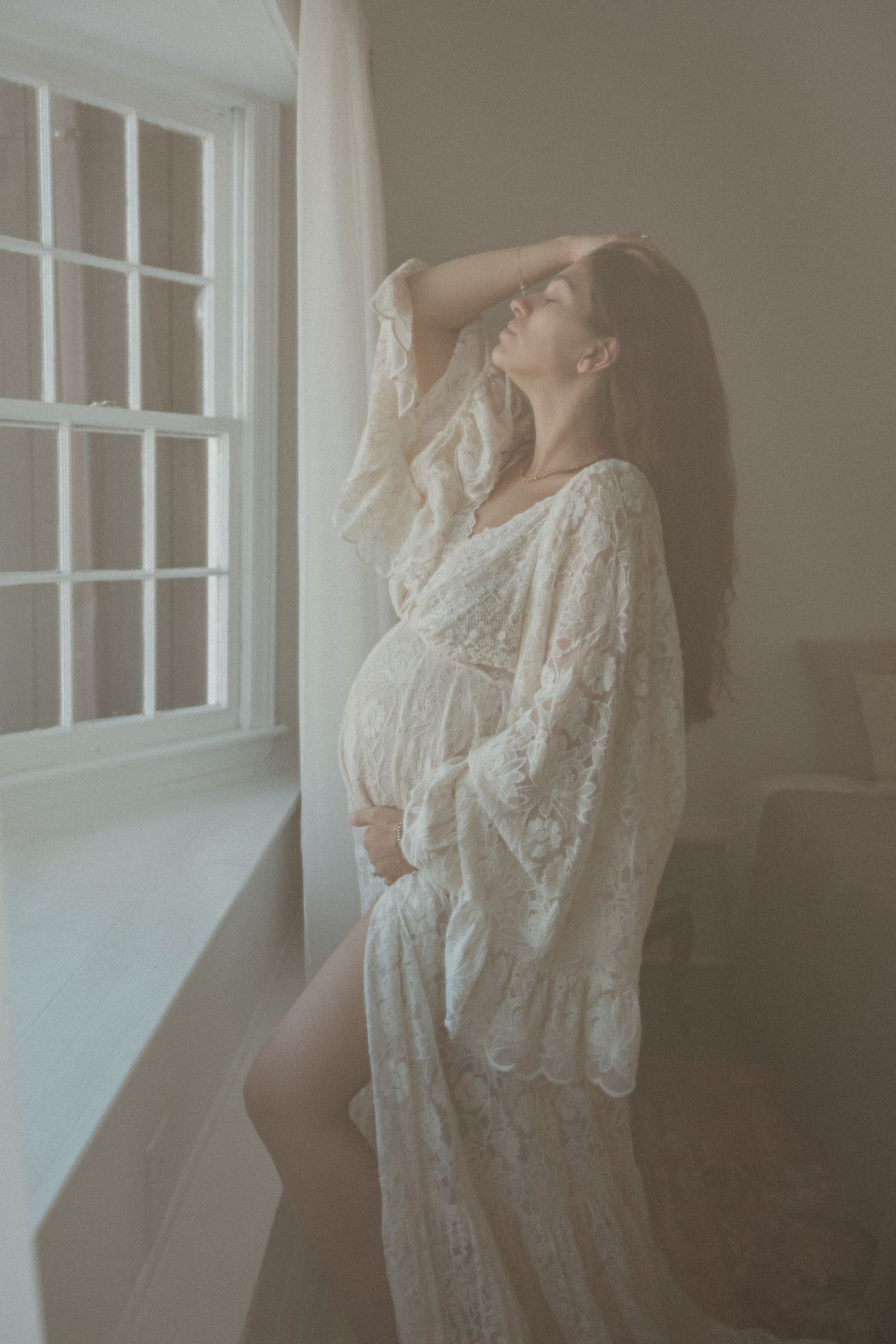 Ethereal maternity photoshoot featuring a delicate white lace robe by a sunlit window.