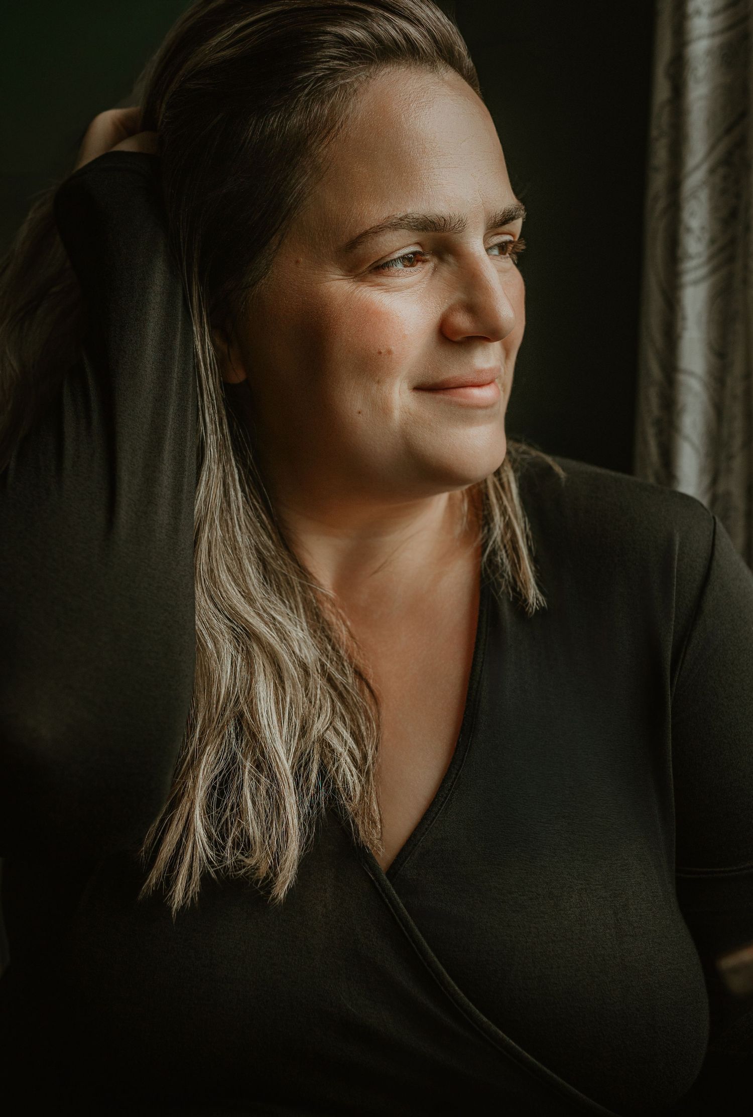 A woman in a black wrap top smiles softly, looking away, with warm light highlighting her face and blonde hair.