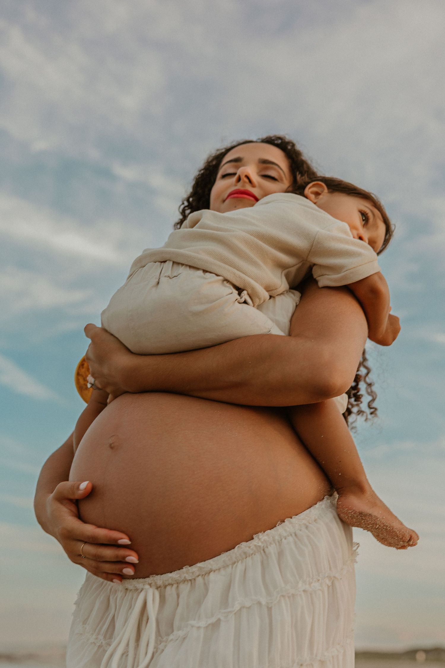 Pregnant woman with curly hair holding a young child outdoors under a cloudy sky.