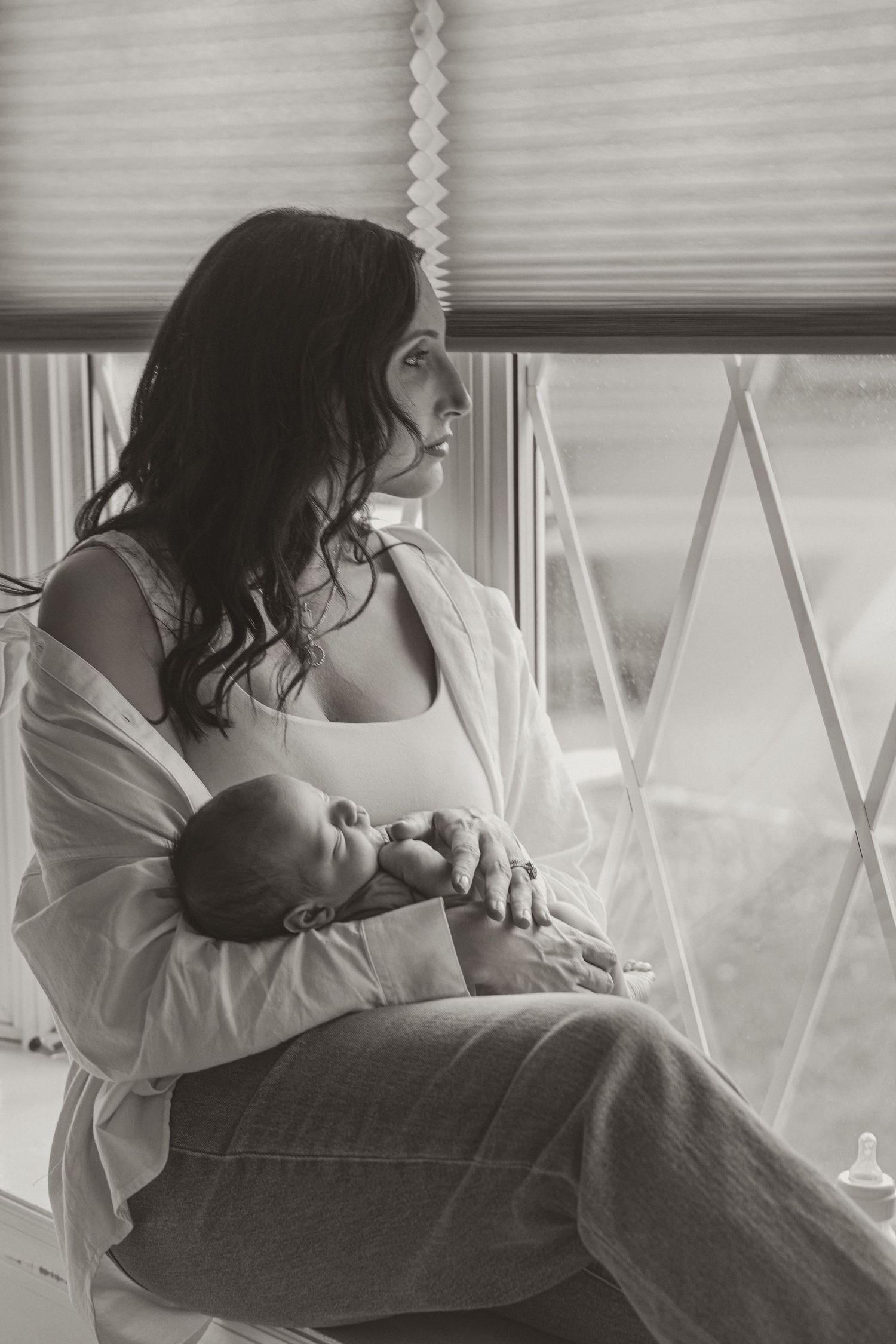 Black and white photo of a young mother sitting by a window, gently cradling her sleeping newborn baby.