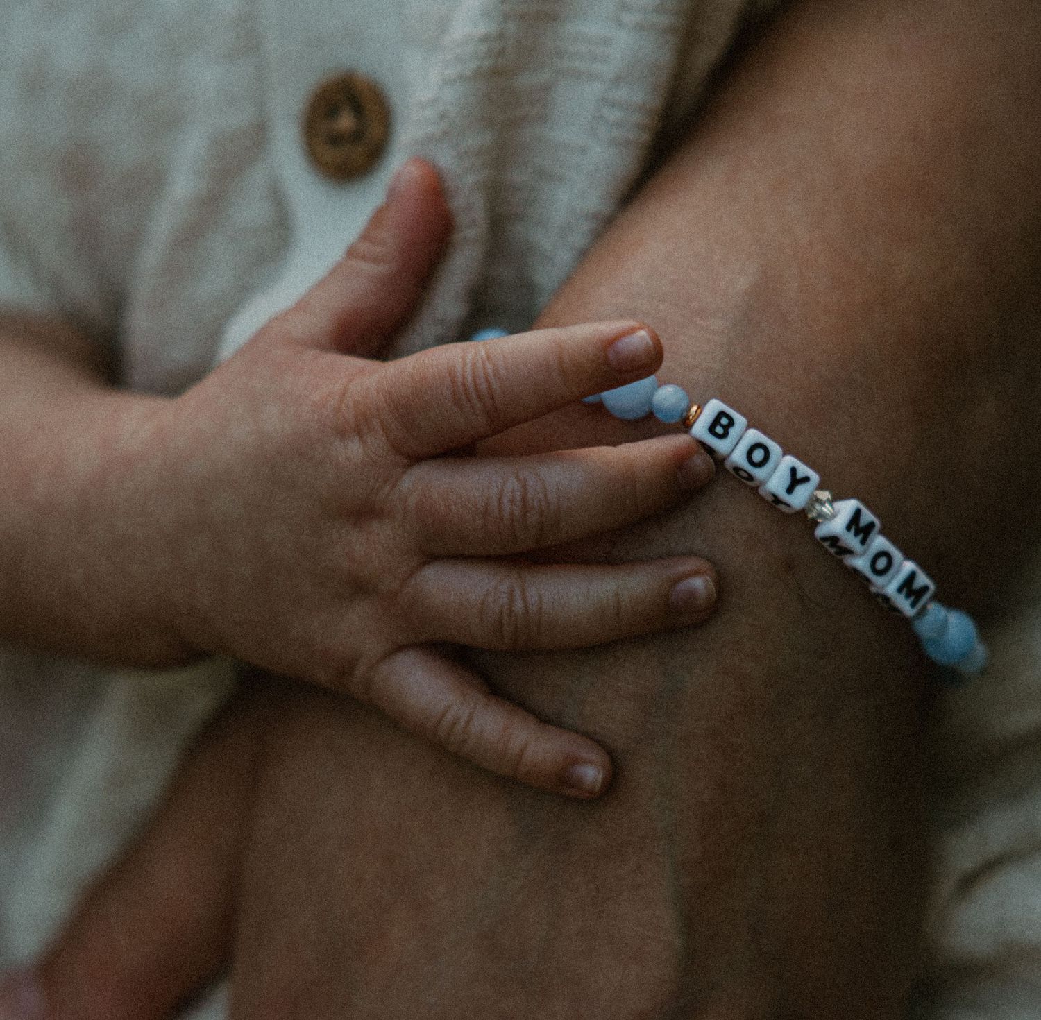 Close-up of a baby hand resting on an adult arm wearing a blue and white beaded BOY MOM bracelet.