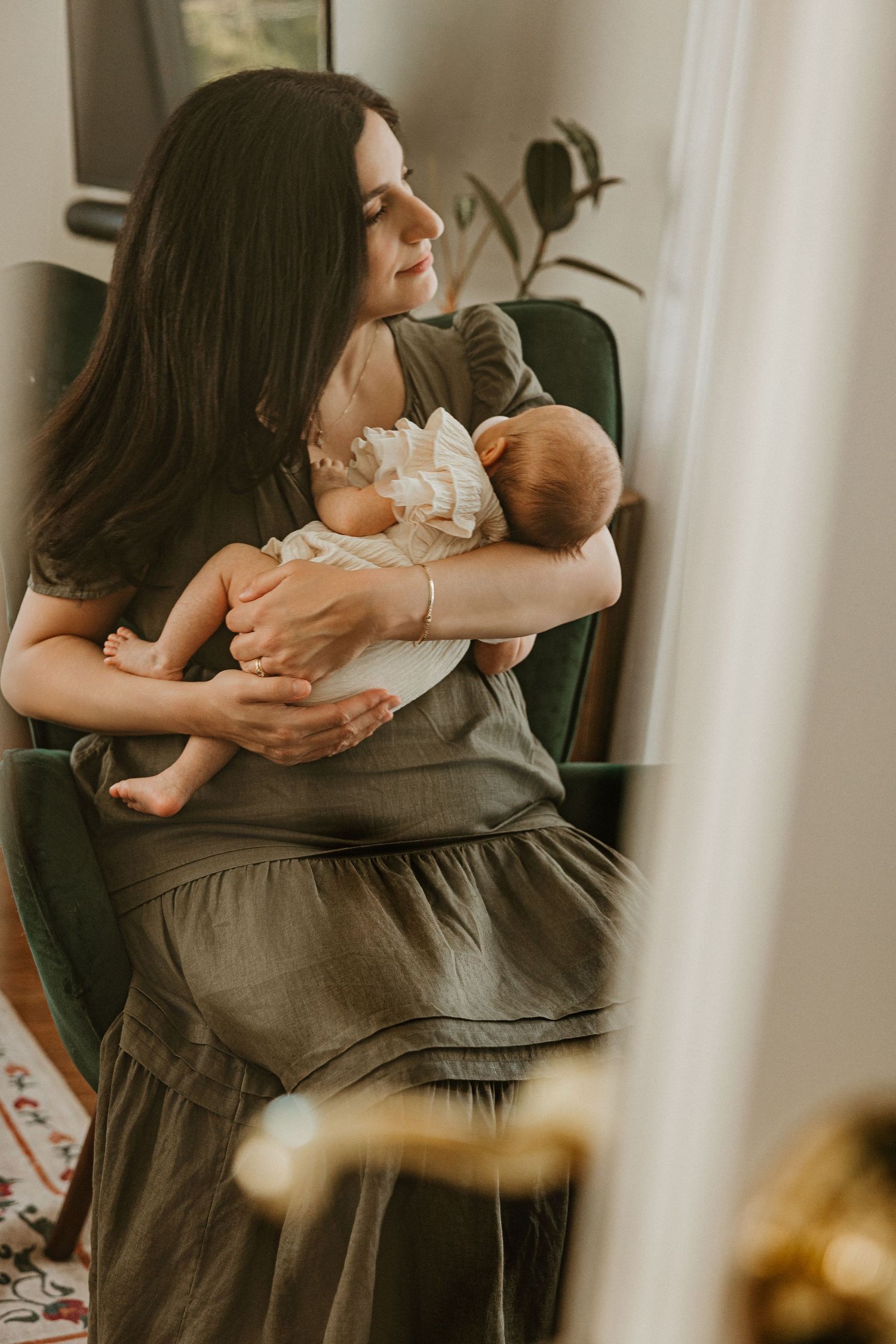 A young mother in an olive dress lovingly gazes at her newborn baby while seated in a green armchair near a window.