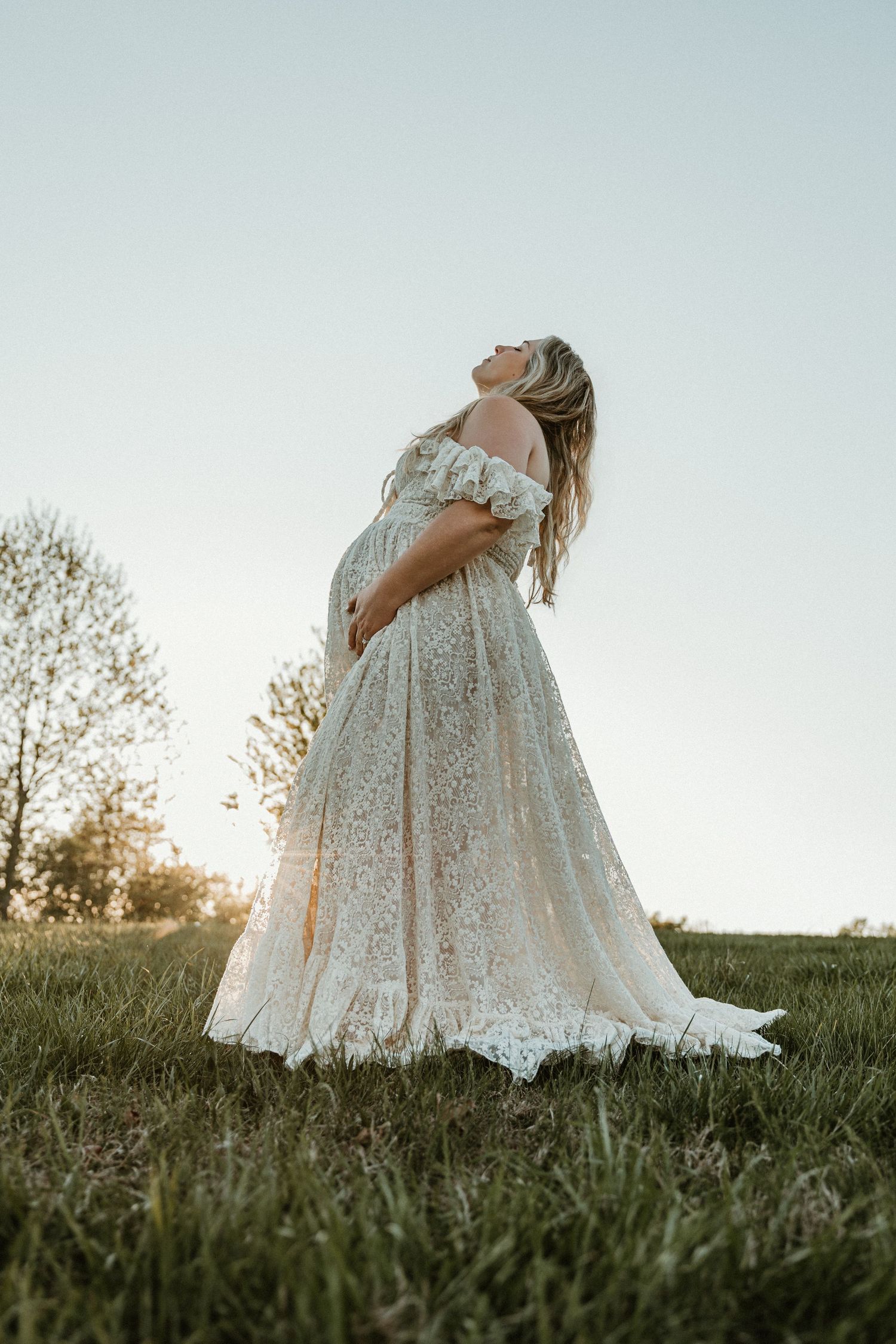 Pregnant woman in off-shoulder lace gown standing in field at golden hour, cradling baby bump while gazing upward.