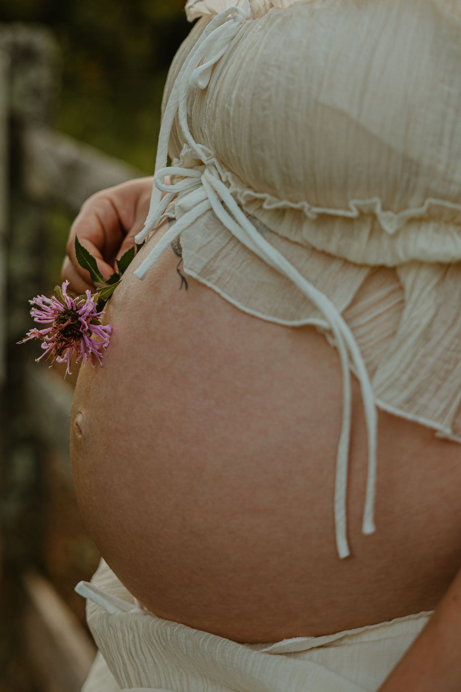 Pregnant woman in white ruffled crop top holding pink wildflowers against her bare baby bump outdoors.