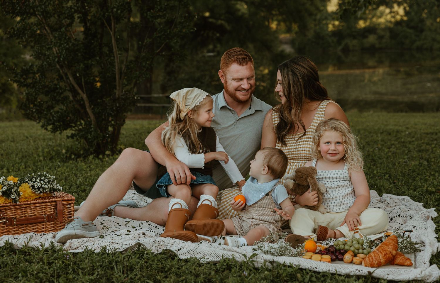 Happy family of five sitting on blanket at outdoor picnic with fruit basket in park.