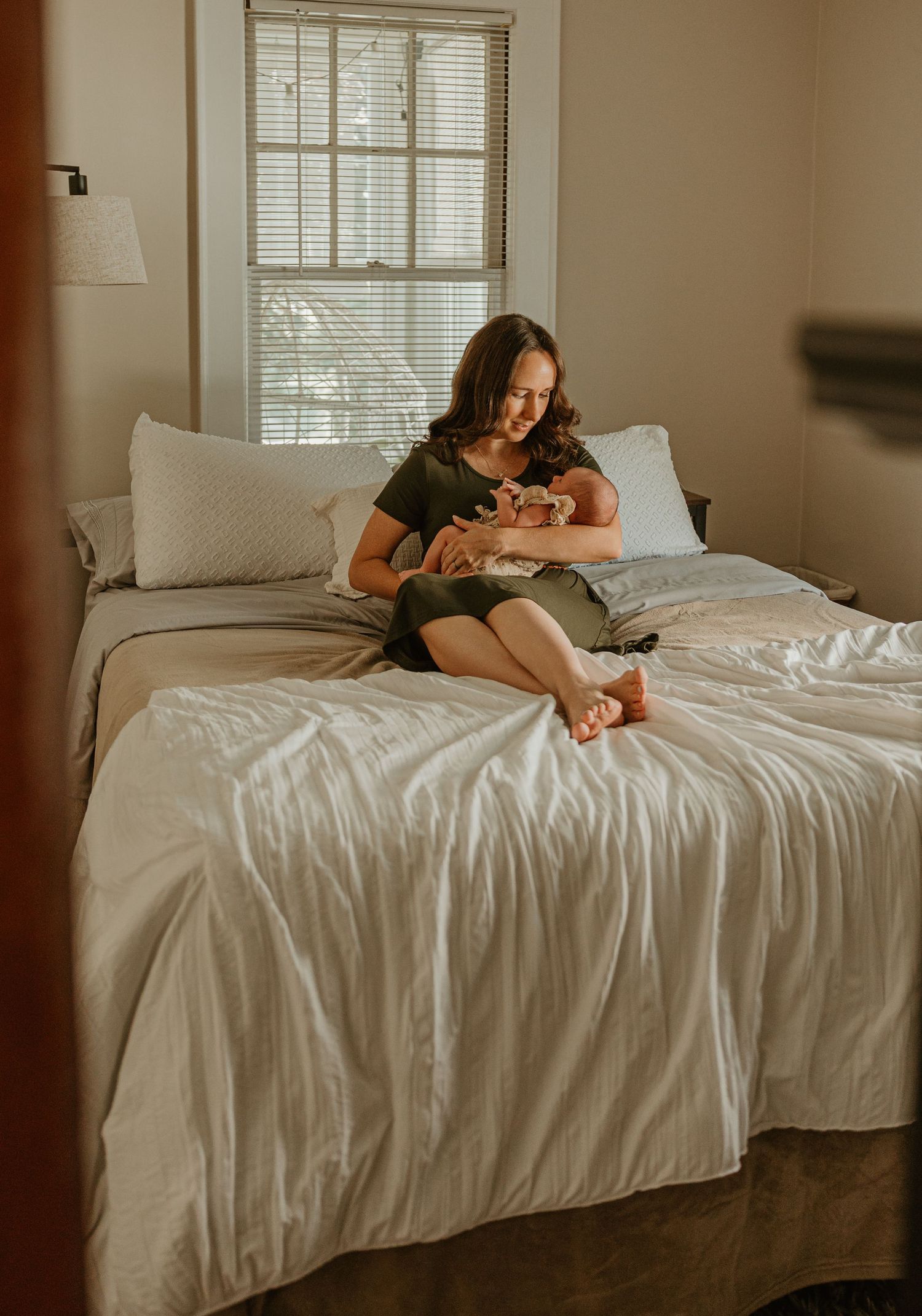 A young mother sits cross-legged on a bed cradling her newborn baby in a softly lit bedroom.