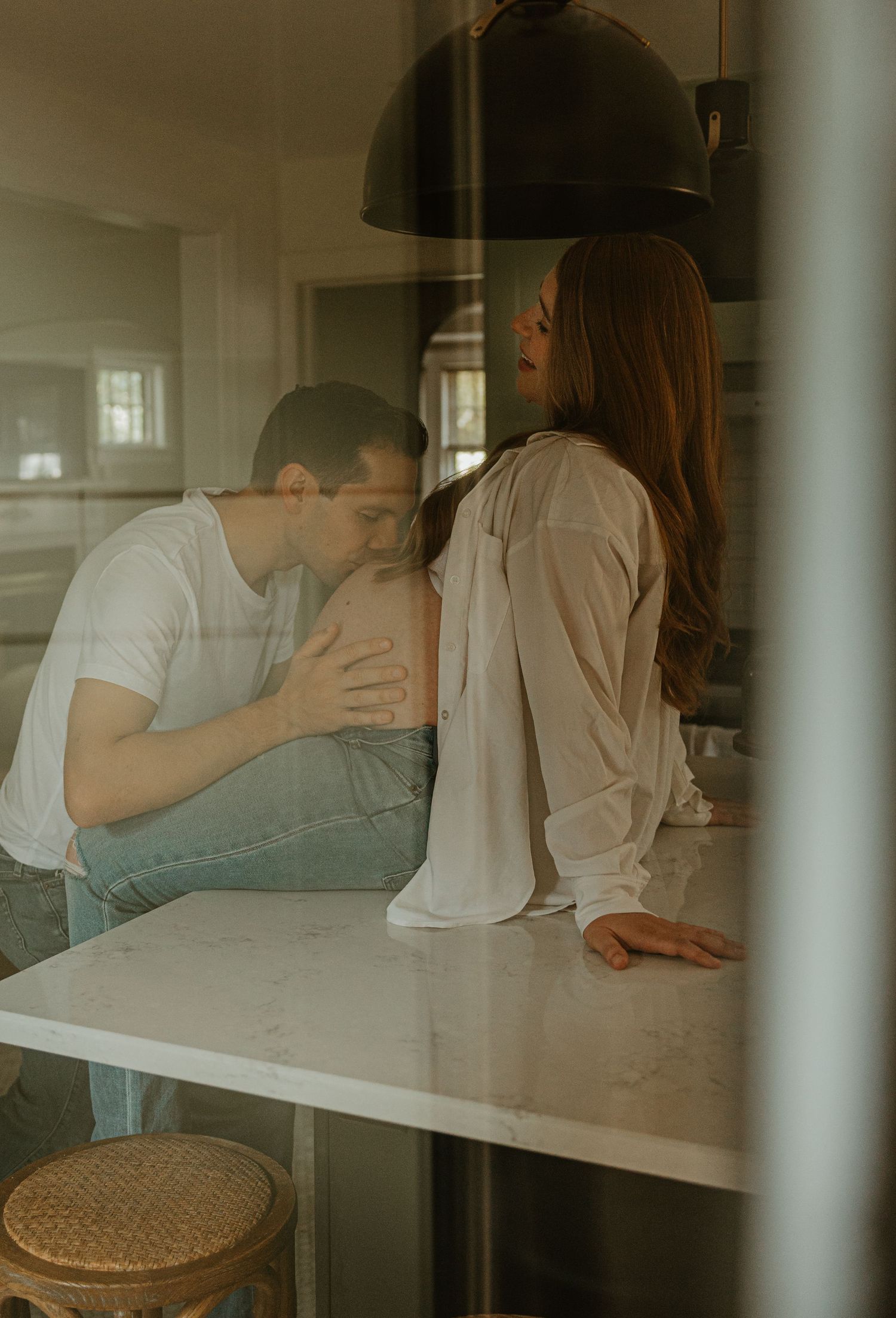 A couple shares an intimate moment in a modern kitchen, the man kissing the woman's shoulder as she leans on a marble counter.