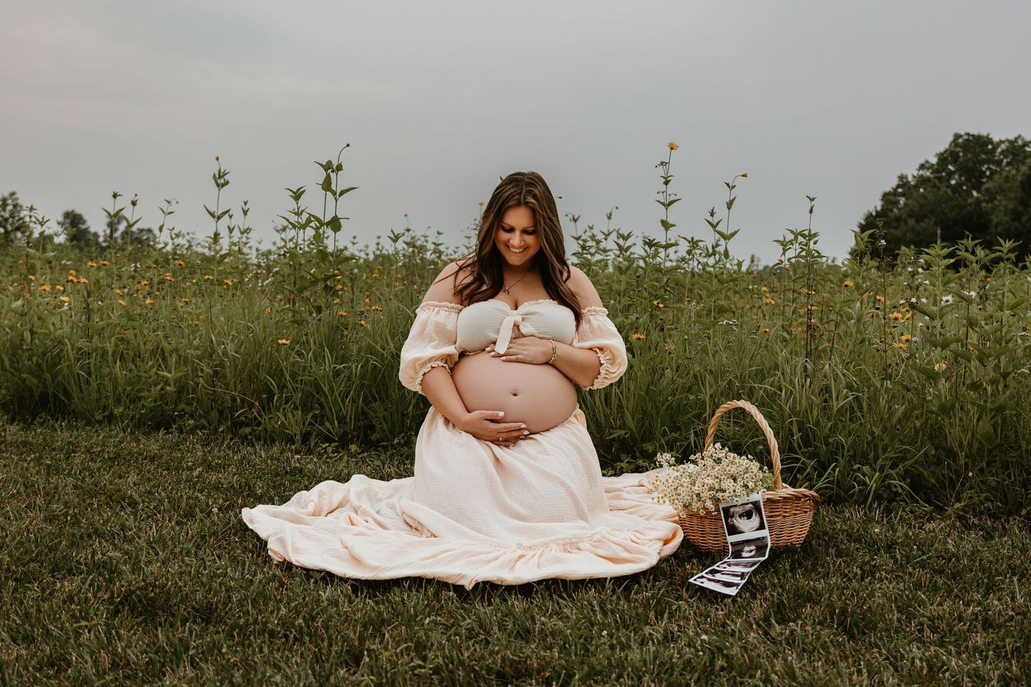 Maternity photoshoot in natural meadow setting with white dress and picnic setup.