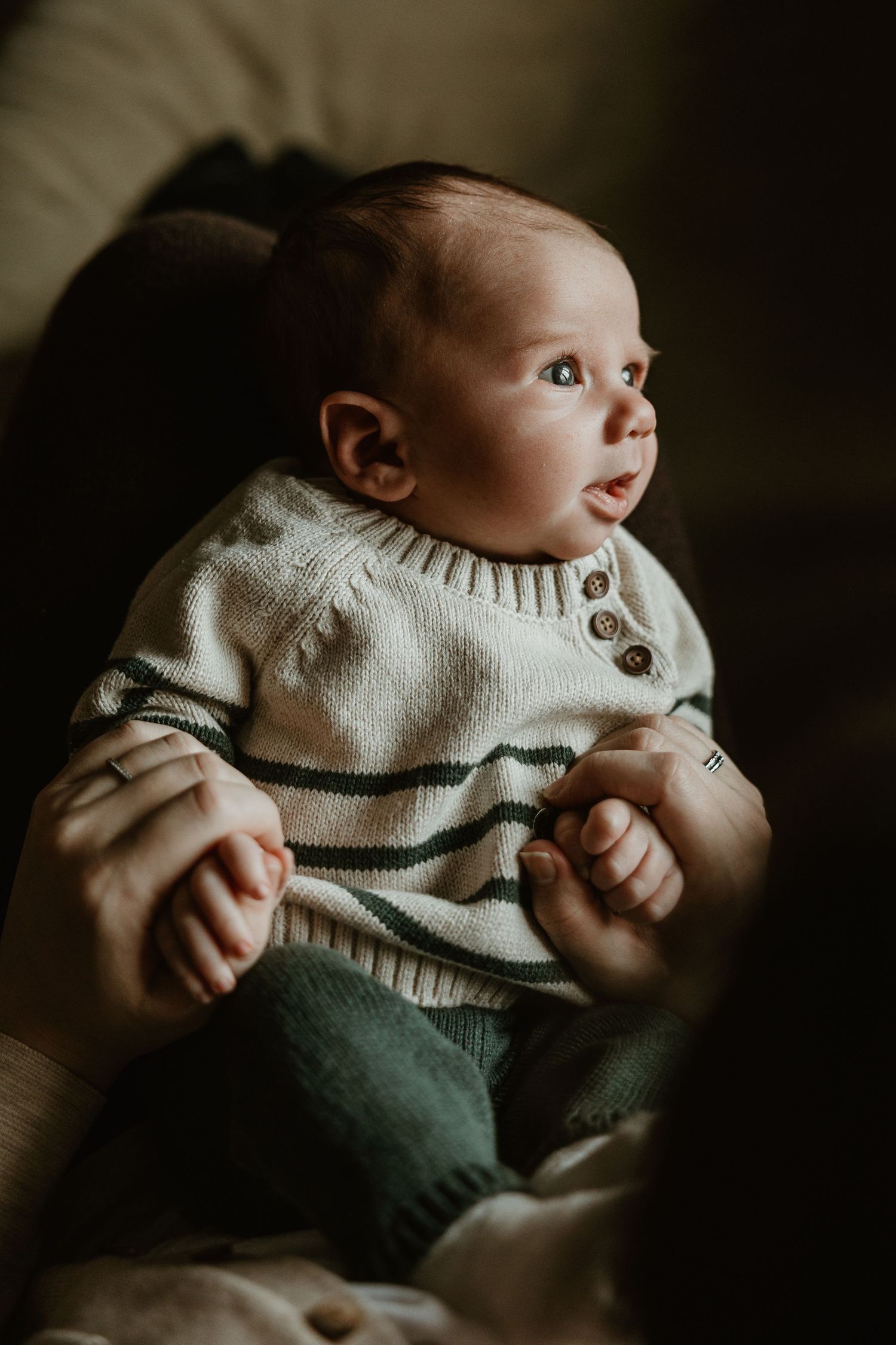 Infant wearing striped sweater gazes upward while being cradled in dark atmospheric setting.