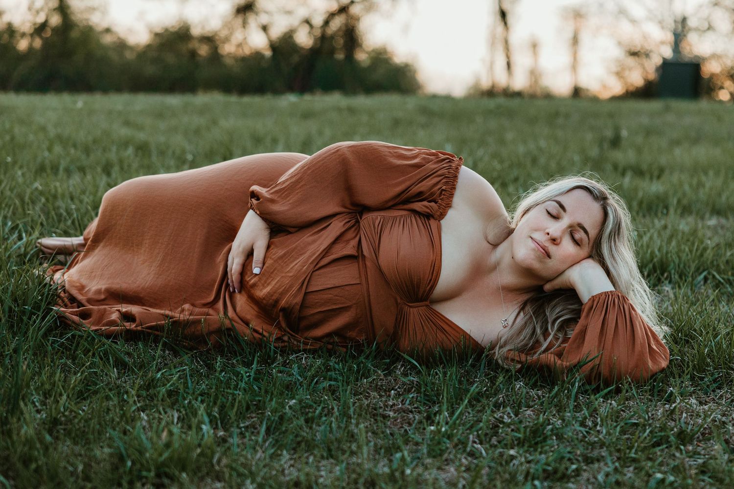 Pregnant woman in rust-orange maternity gown lying on green grass during golden hour outdoor photoshoot.