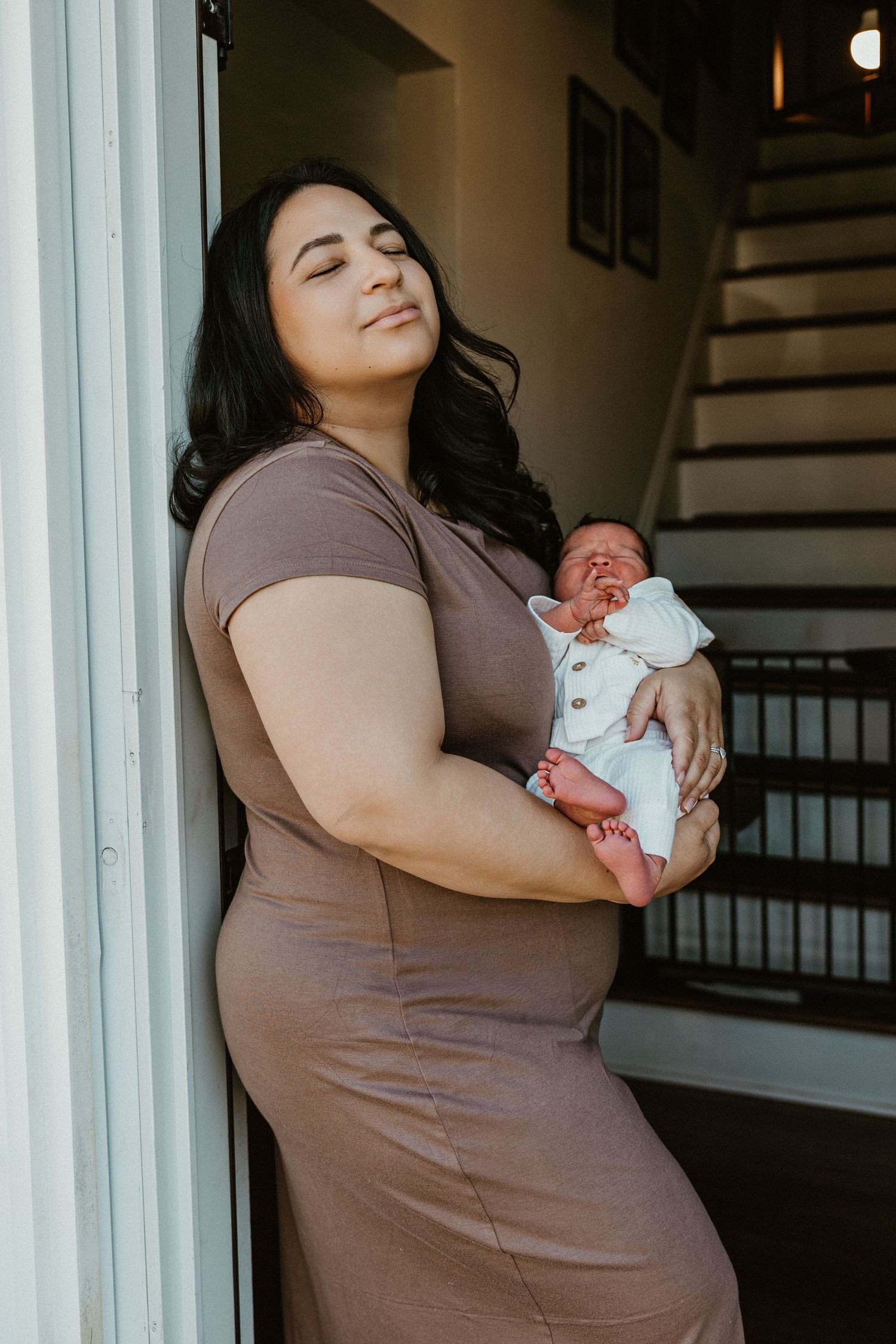 A woman in a brown dress holds a newborn baby while leaning against a doorframe with eyes closed.