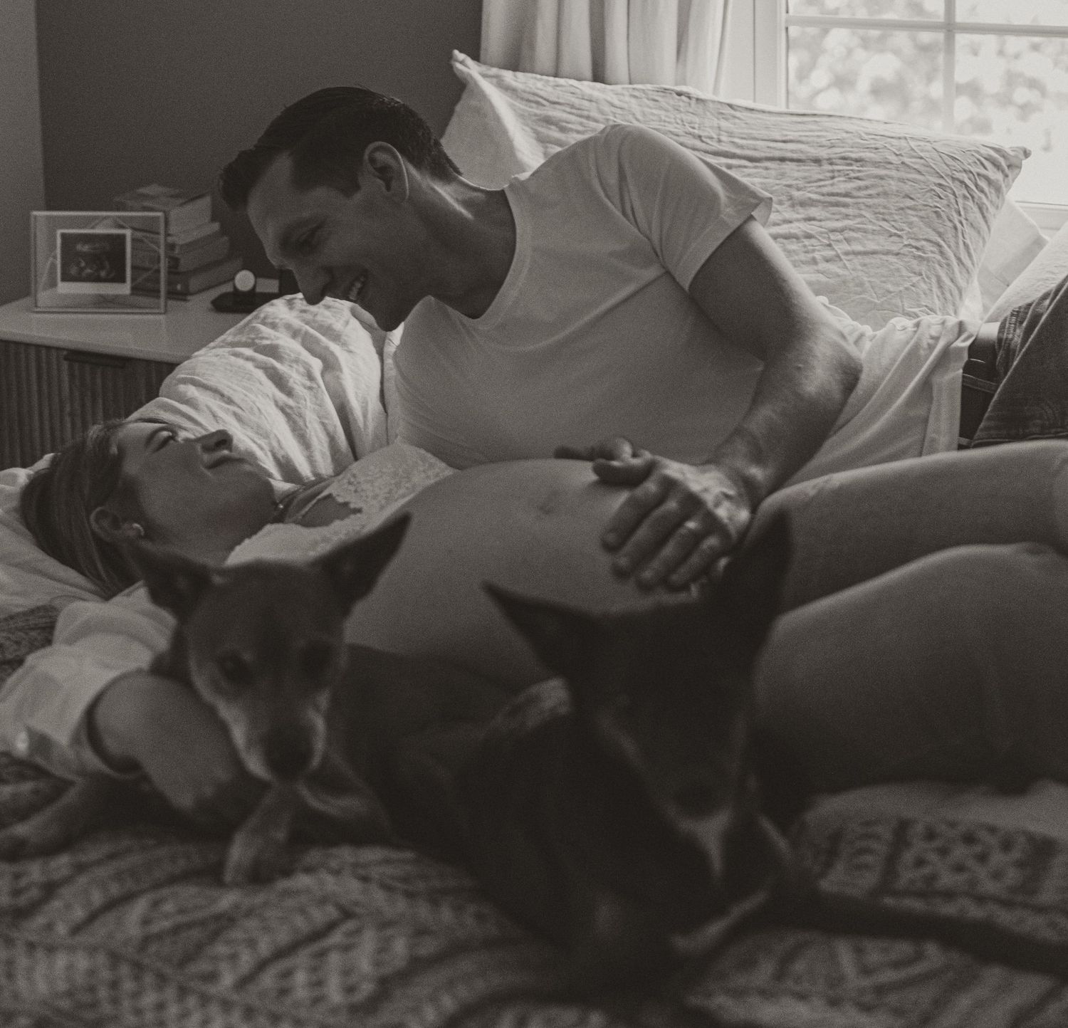 Black and white photo of a couple sharing a tender moment on a bed with their dog nearby.