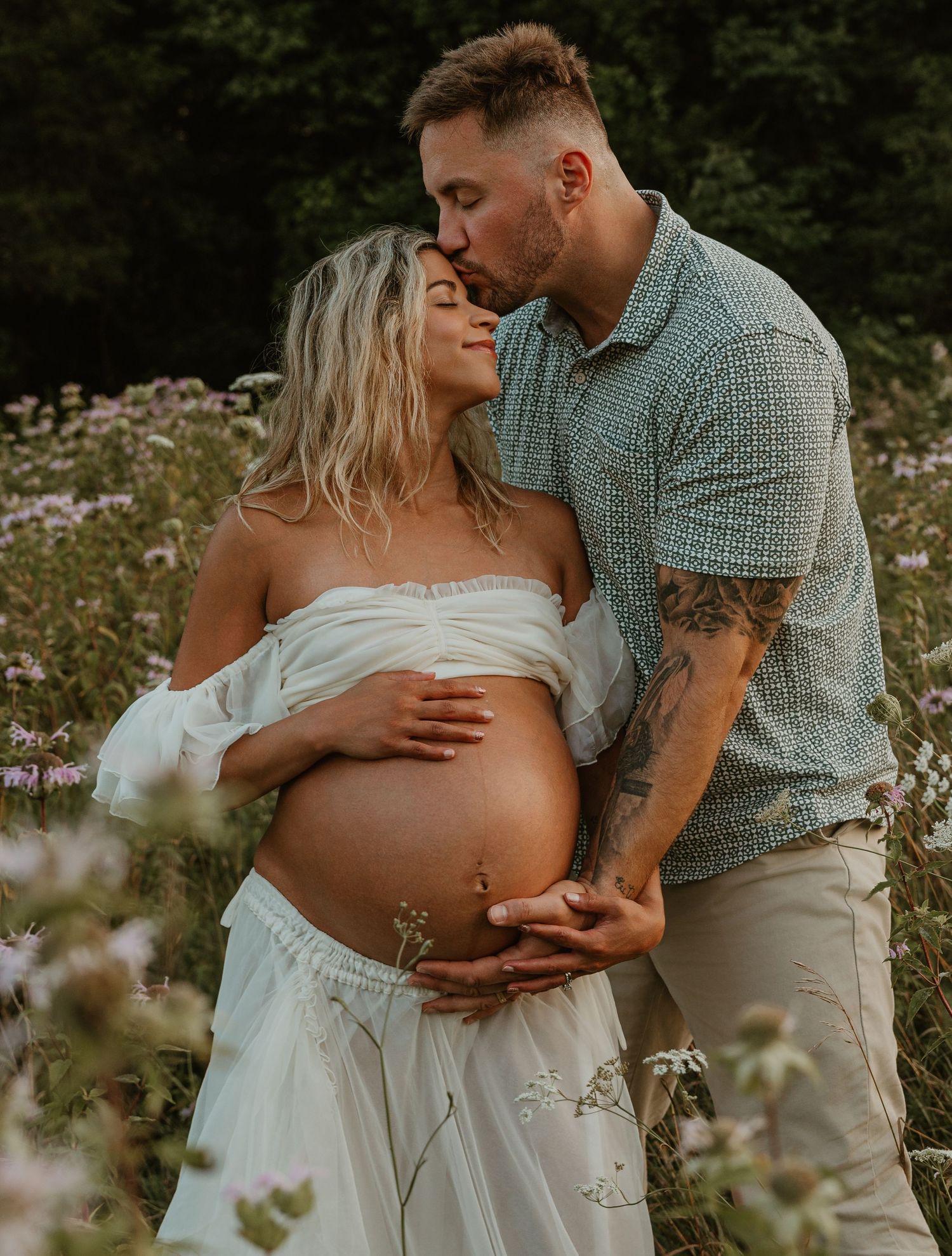 A man kisses a pregnant woman in a white dress while embracing her baby bump in a wildflower field.