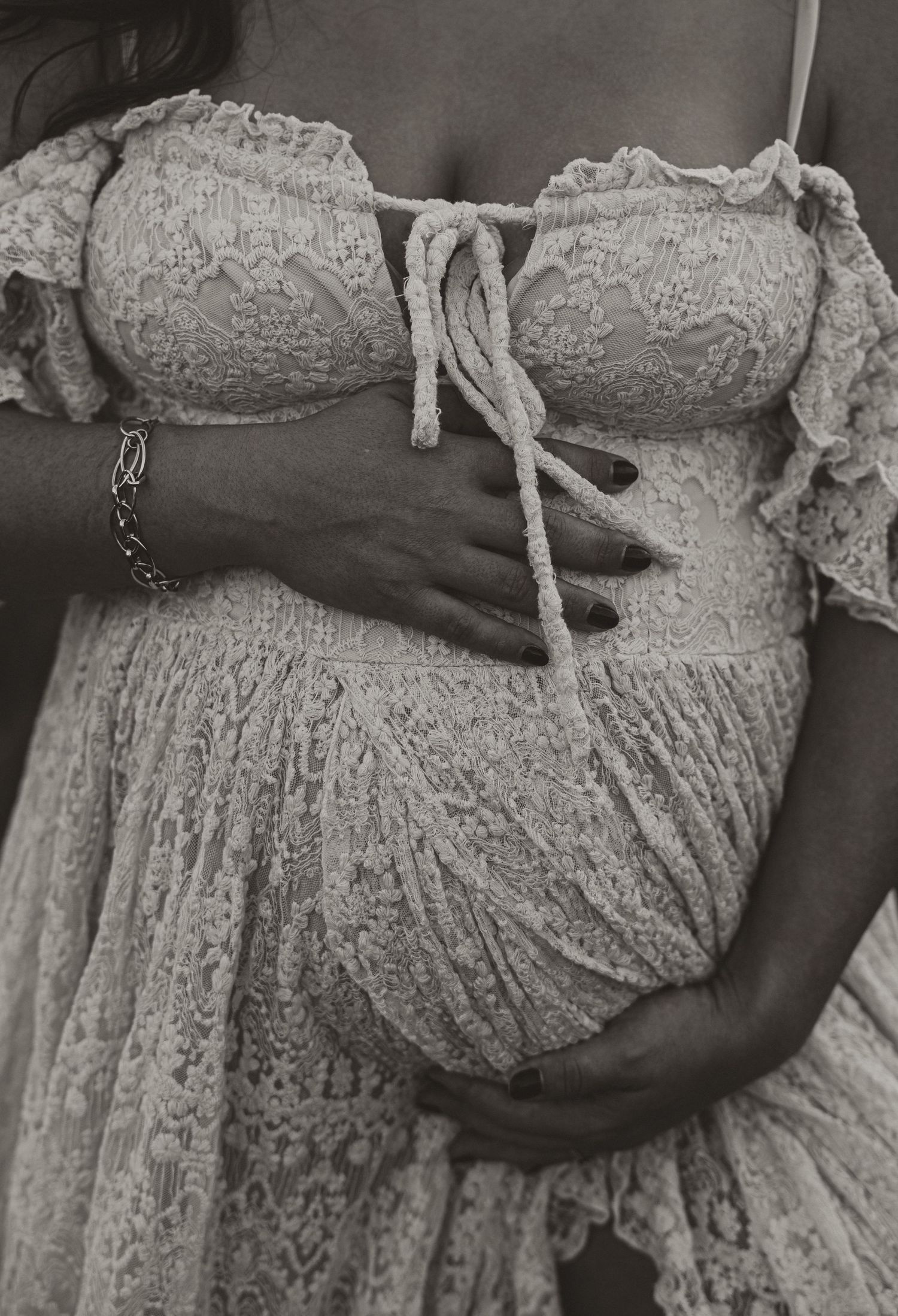 Black and white photo of a pregnant woman in lace dress cradling her baby bump with both hands.