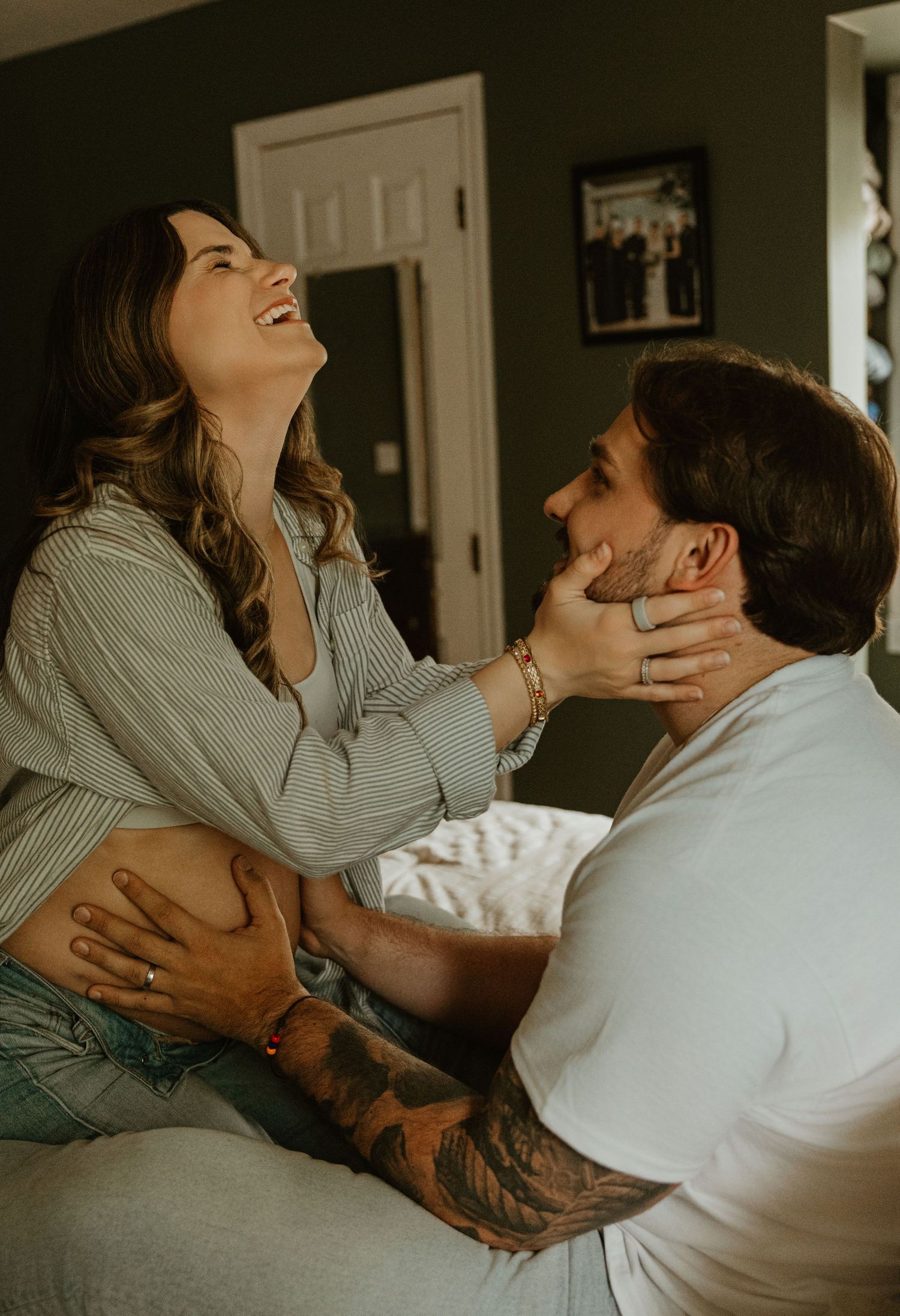 A laughing pregnant woman holds a tattooed man's face while sitting together on a bed in a cozy home.