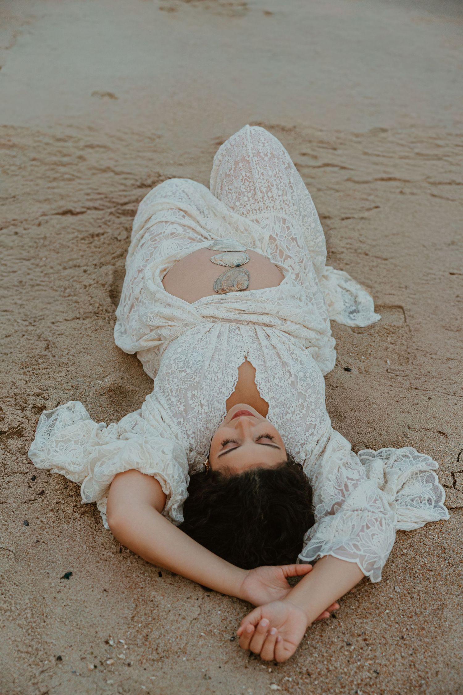 Woman in white lace dress lying peacefully on sandy beach with eyes closed.