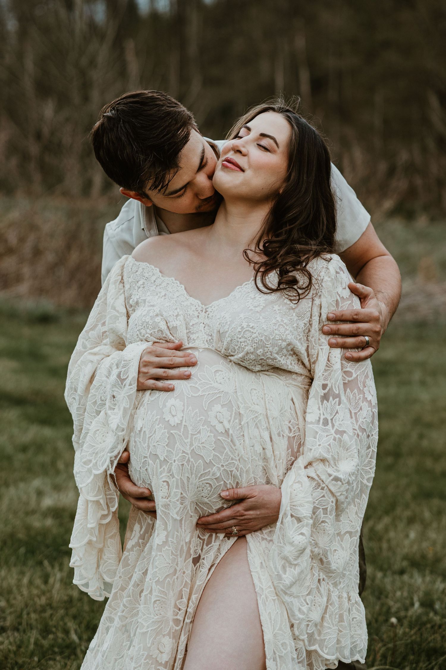 Couple sharing tender maternity photo outdoors, pregnant woman in lace boho dress cradled by partner in green field.