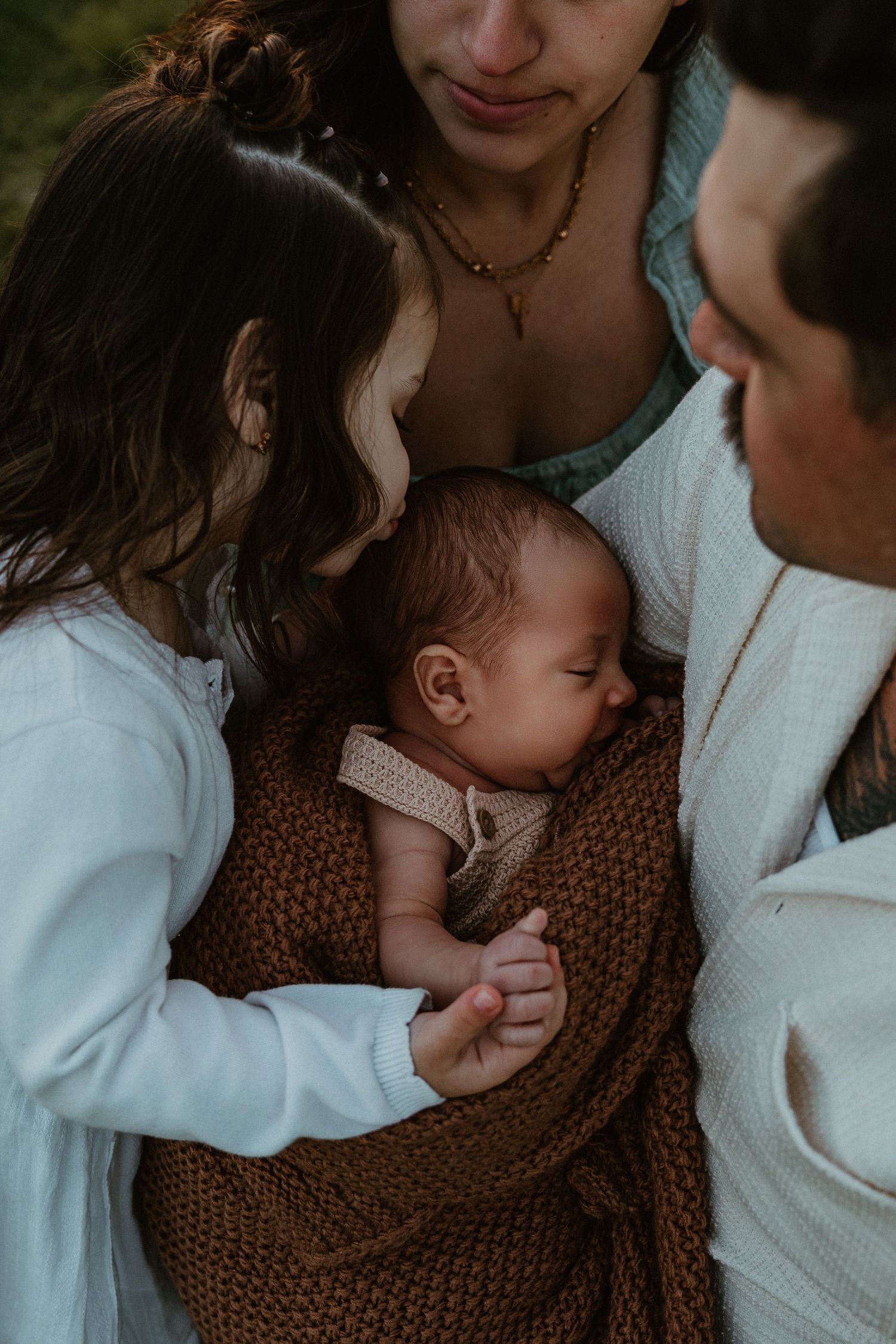 Family gathered around a sleeping newborn wrapped in a brown knit blanket, sharing a tender moment outdoors.