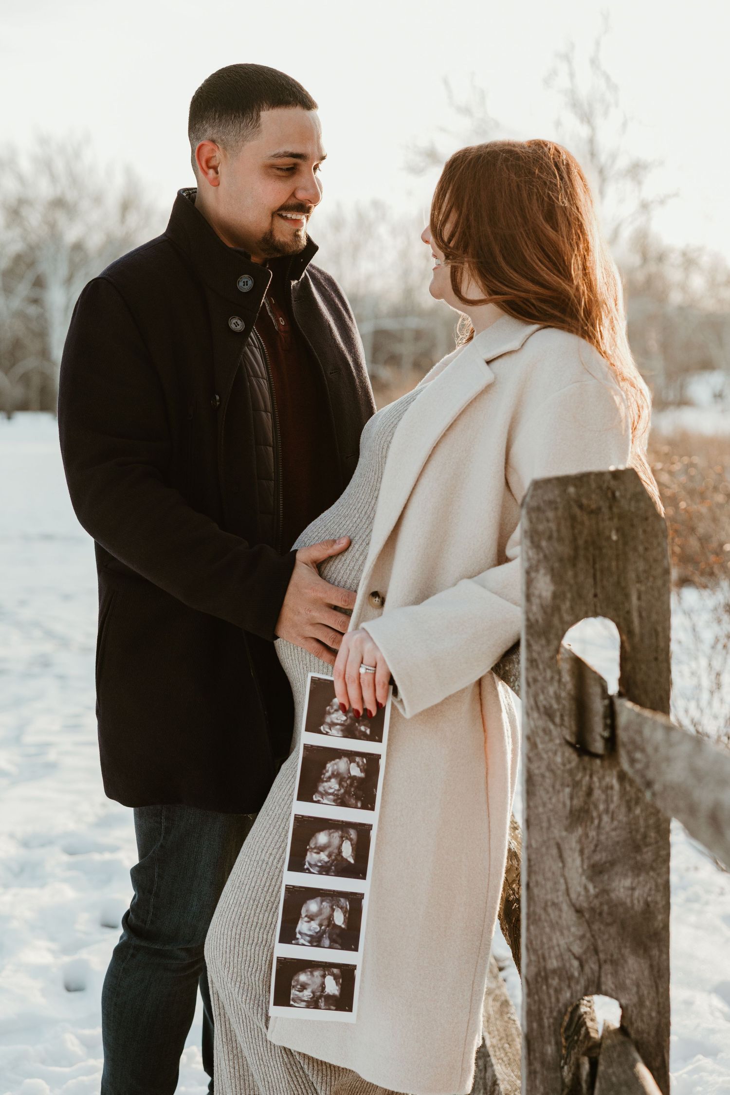 Couple holds ultrasound photos in snowy winter setting during pregnancy announcement shoot.