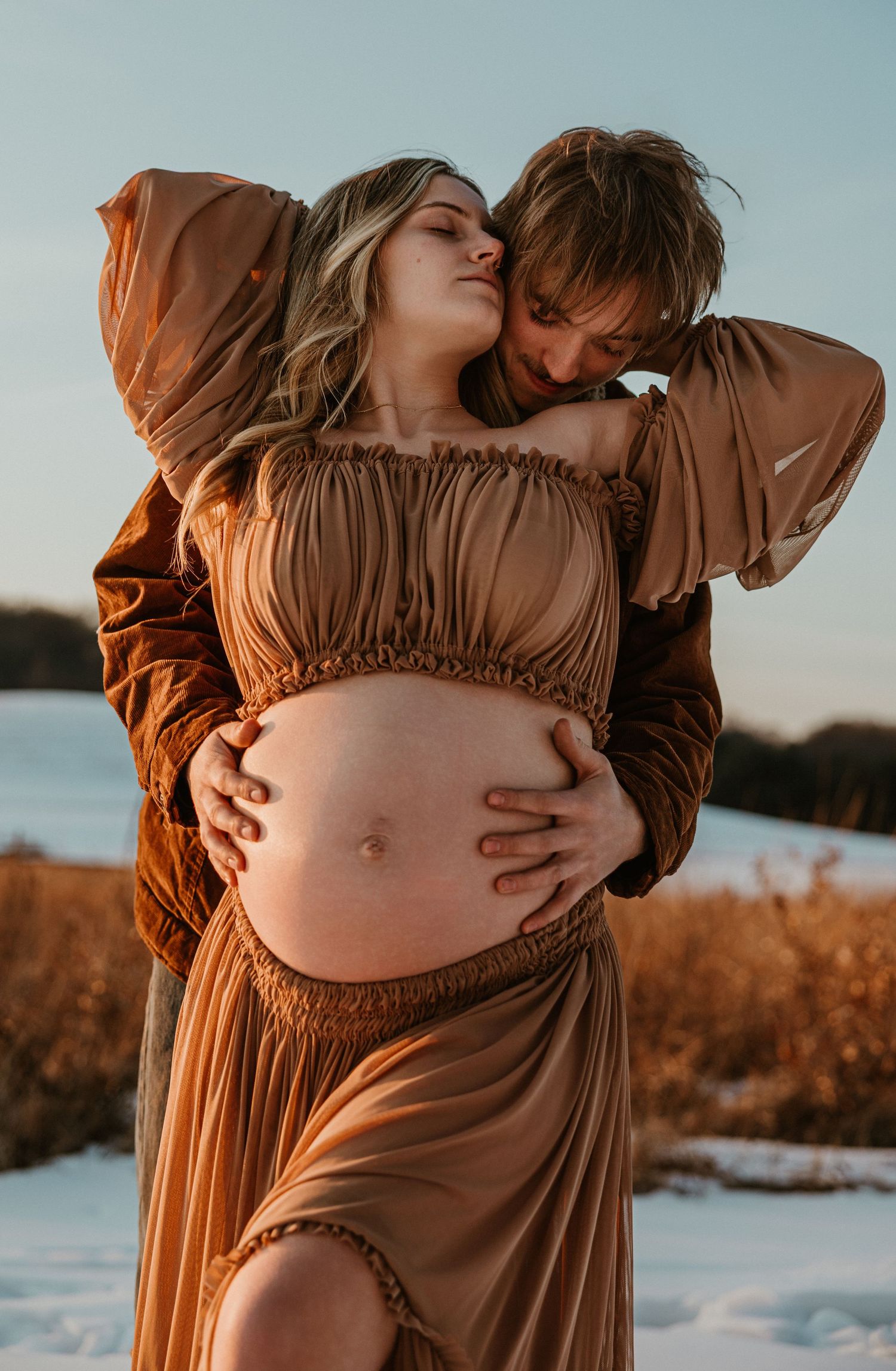 Intimate maternity photos at sunset showcase flowing brown dress against snowy backdrop.