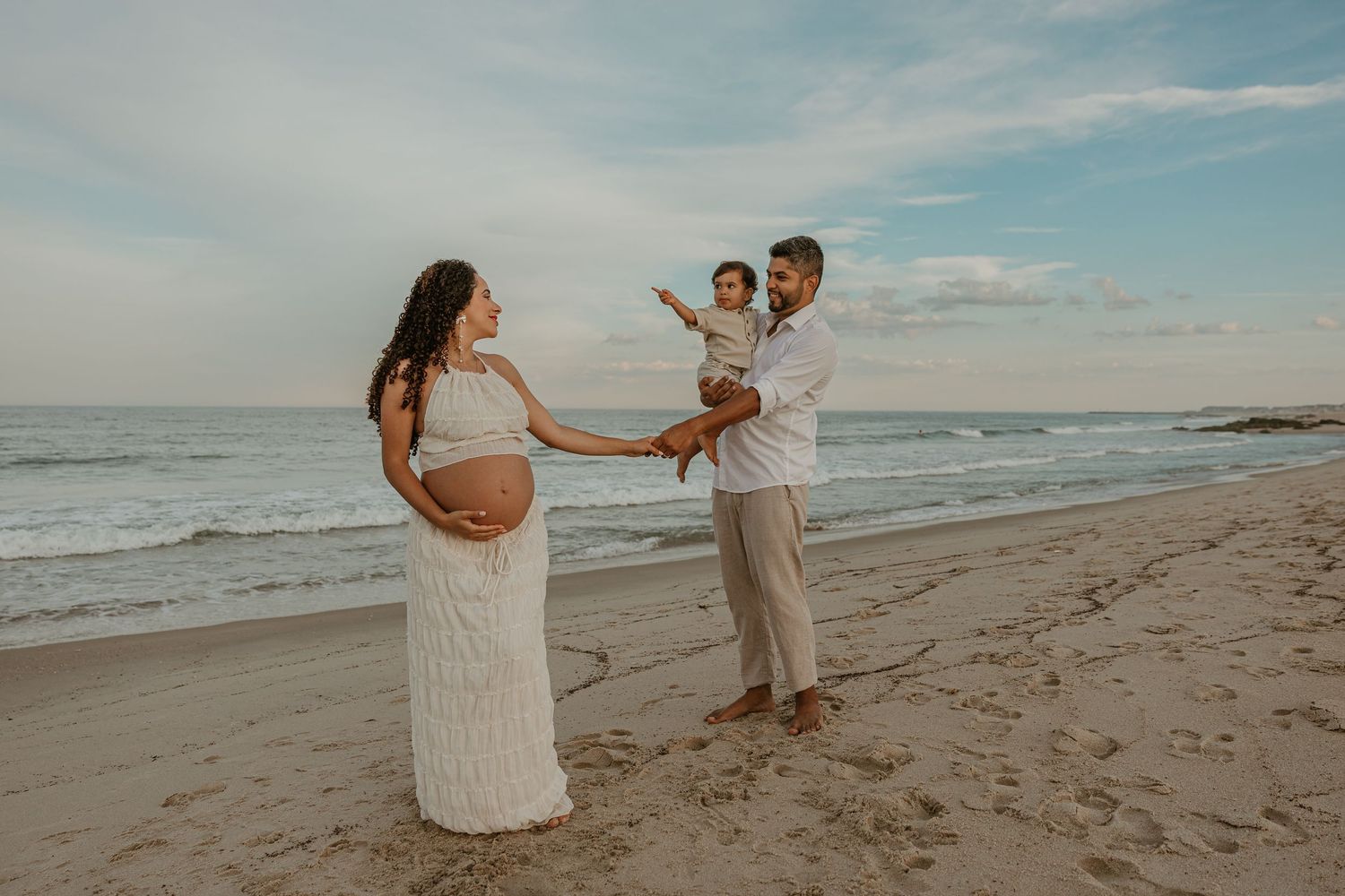 Pregnant woman in white lace gown with husband holding toddler on sandy beach during maternity photo shoot.