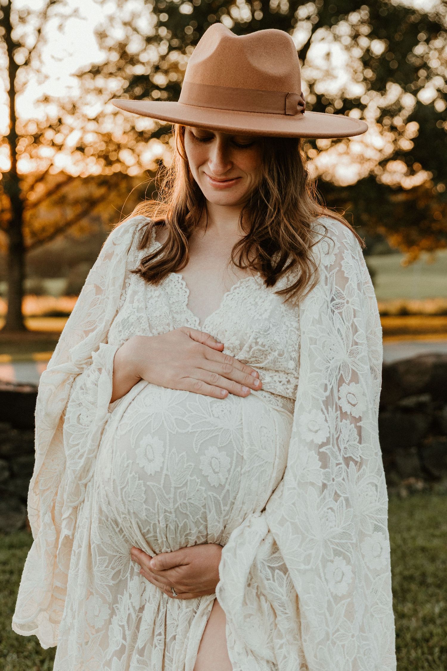 Maternity portrait with brown hat and white lace dress during golden hour.