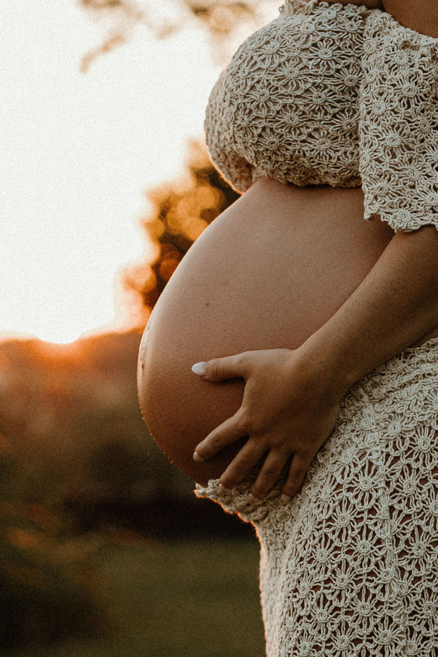 Close-up side view of pregnant belly in white lace dress at sunset.