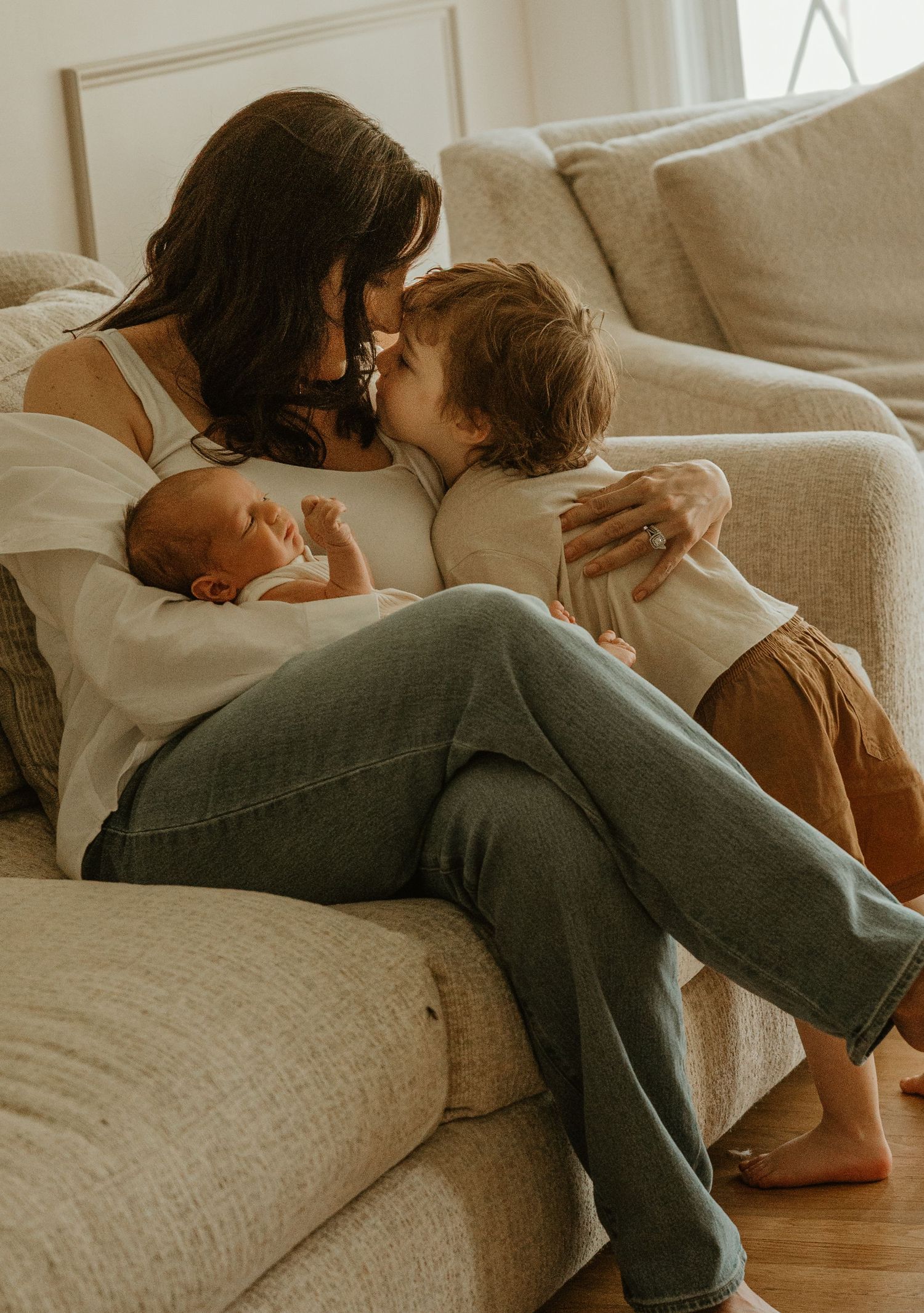 Mother sits on couch kissing toddler while cradling newborn baby in warm cozy living room.