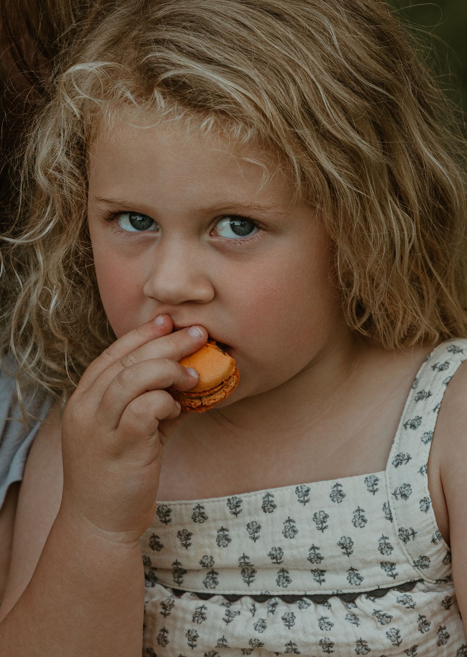 Young curly-haired girl with blue eyes eating an orange macaron cookie, wearing a floral sundress.