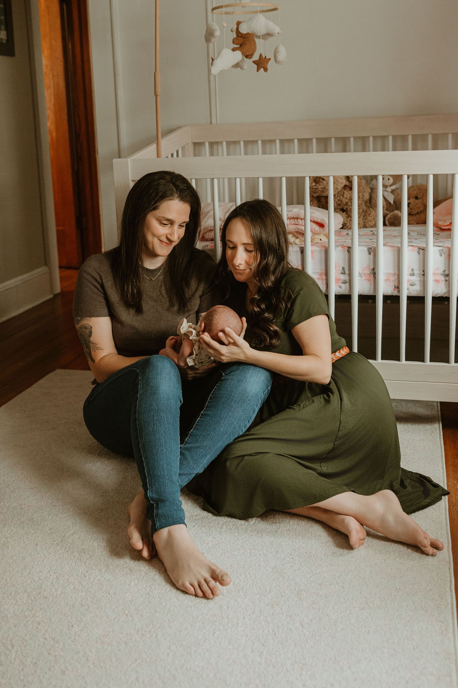 Two mothers sitting on nursery floor holding newborn baby near white crib in cozy bedroom.