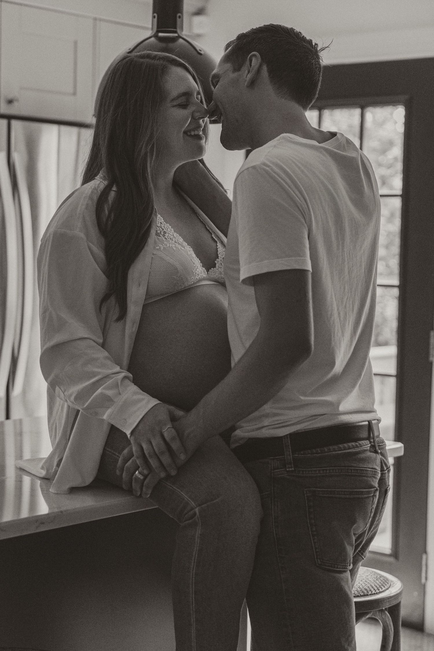 Black and white photo of a smiling pregnant couple sharing an intimate moment together in a cozy kitchen setting.