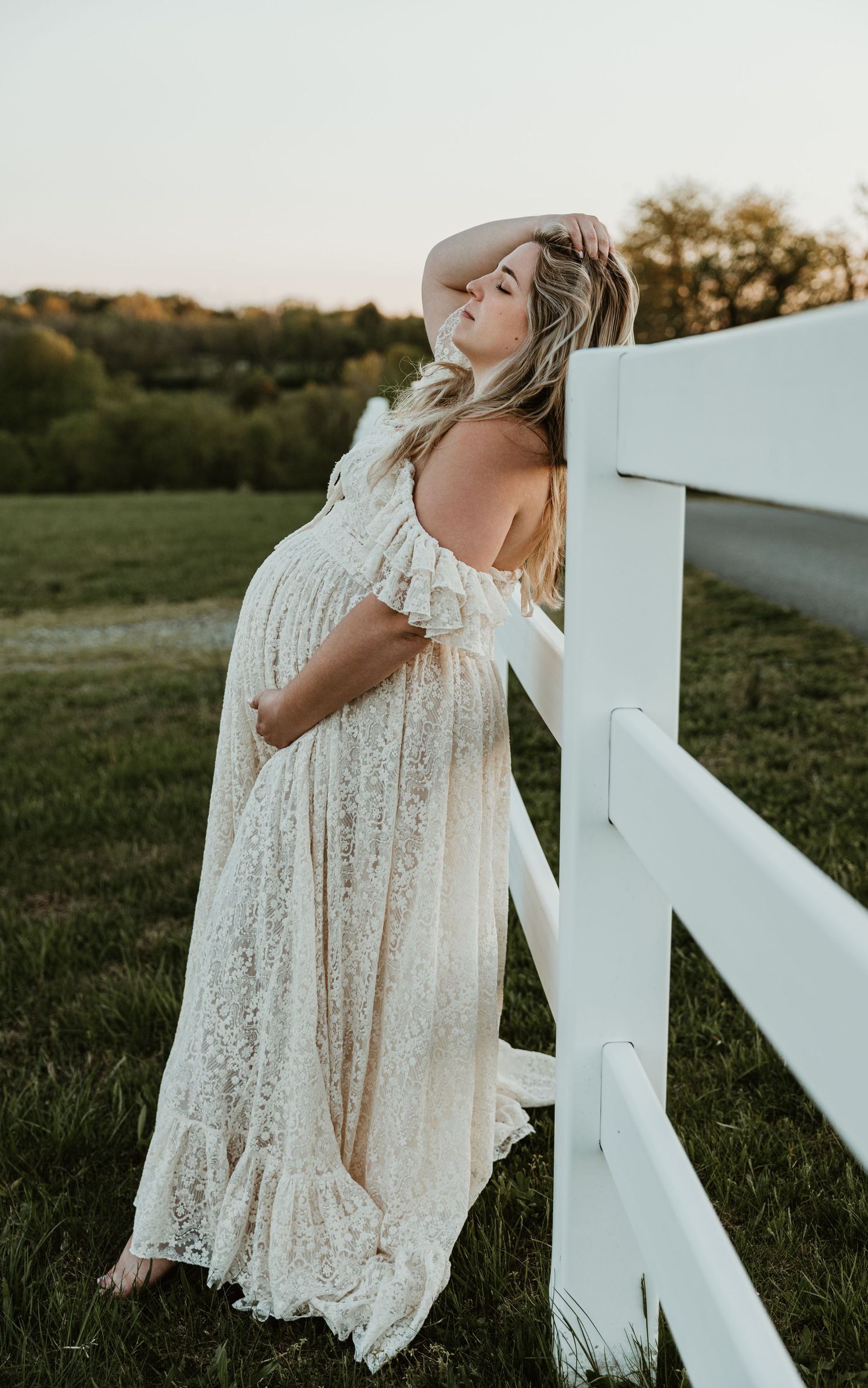 Pregnant woman in ivory lace gown leaning against white fence at golden hour in a green field.