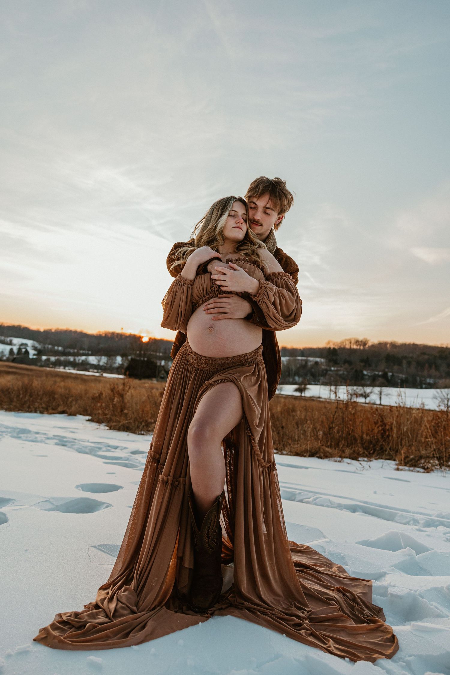 Winter maternity photo of couple embracing in brown dress against snowy landscape at sunset.