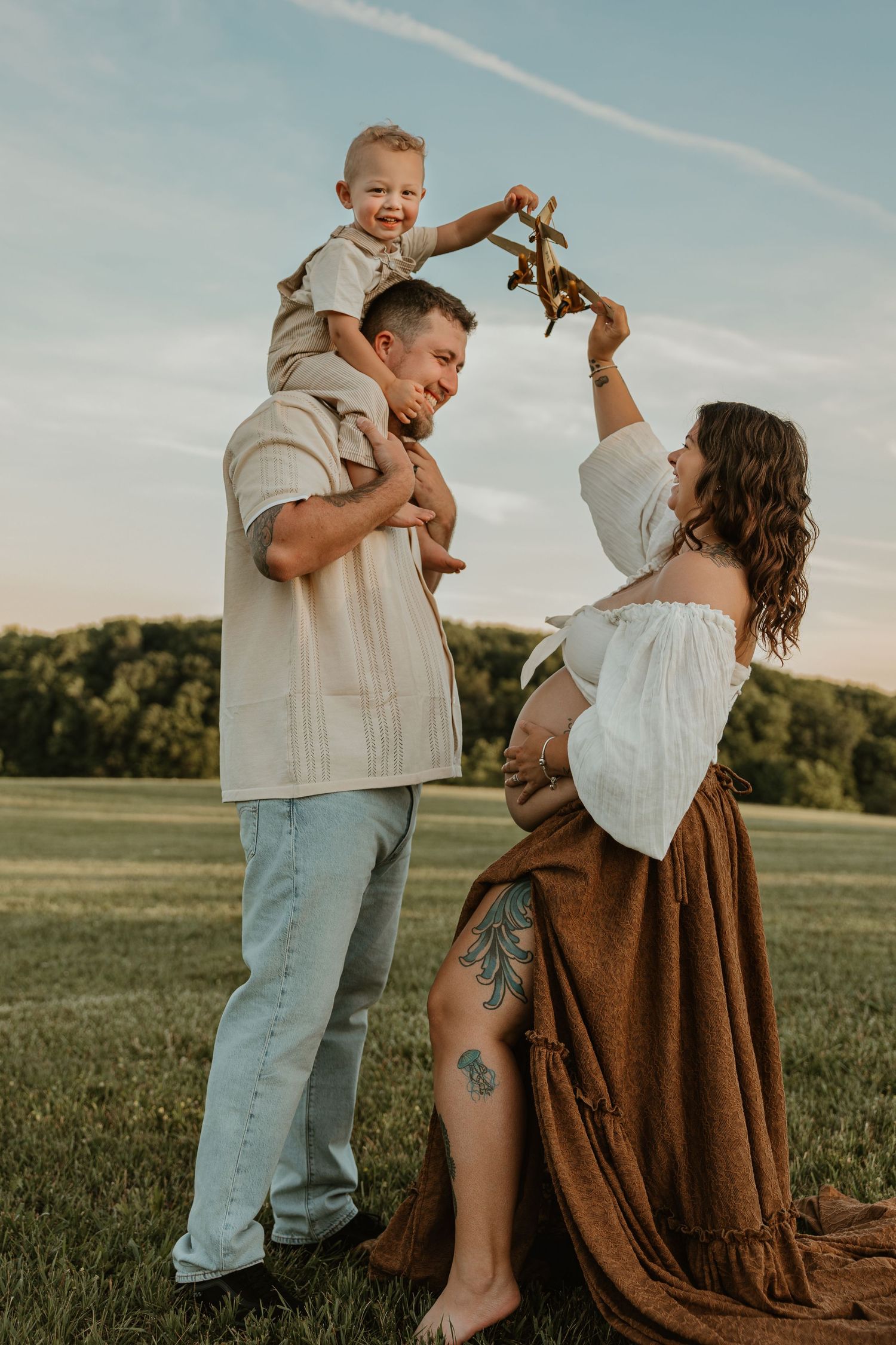 Pregnant woman in white off-shoulder top and brown skirt with tattooed leg plays with toddler on man's shoulders outdoors.