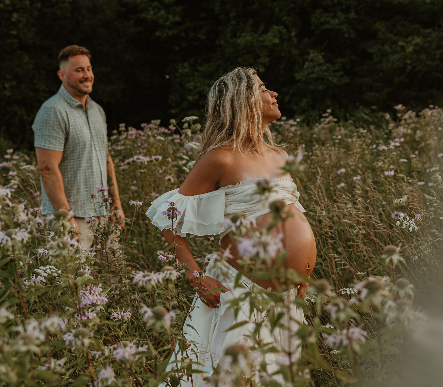 Pregnant woman in white off-shoulder dress stands in wildflower field with smiling man during outdoor maternity photo session.