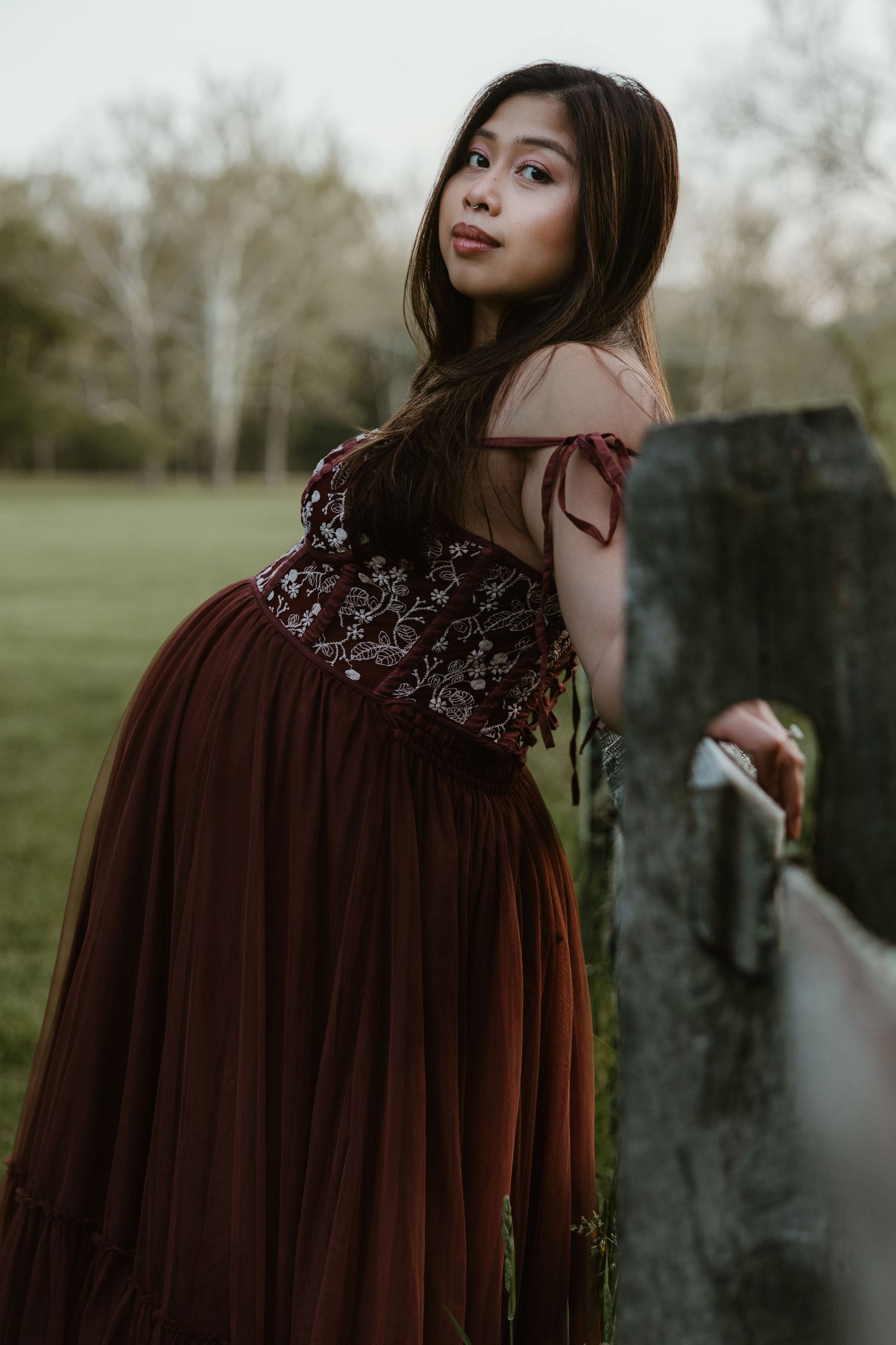 Young woman in elegant burgundy off-shoulder gown poses gracefully by a rustic fence in a lush green park.