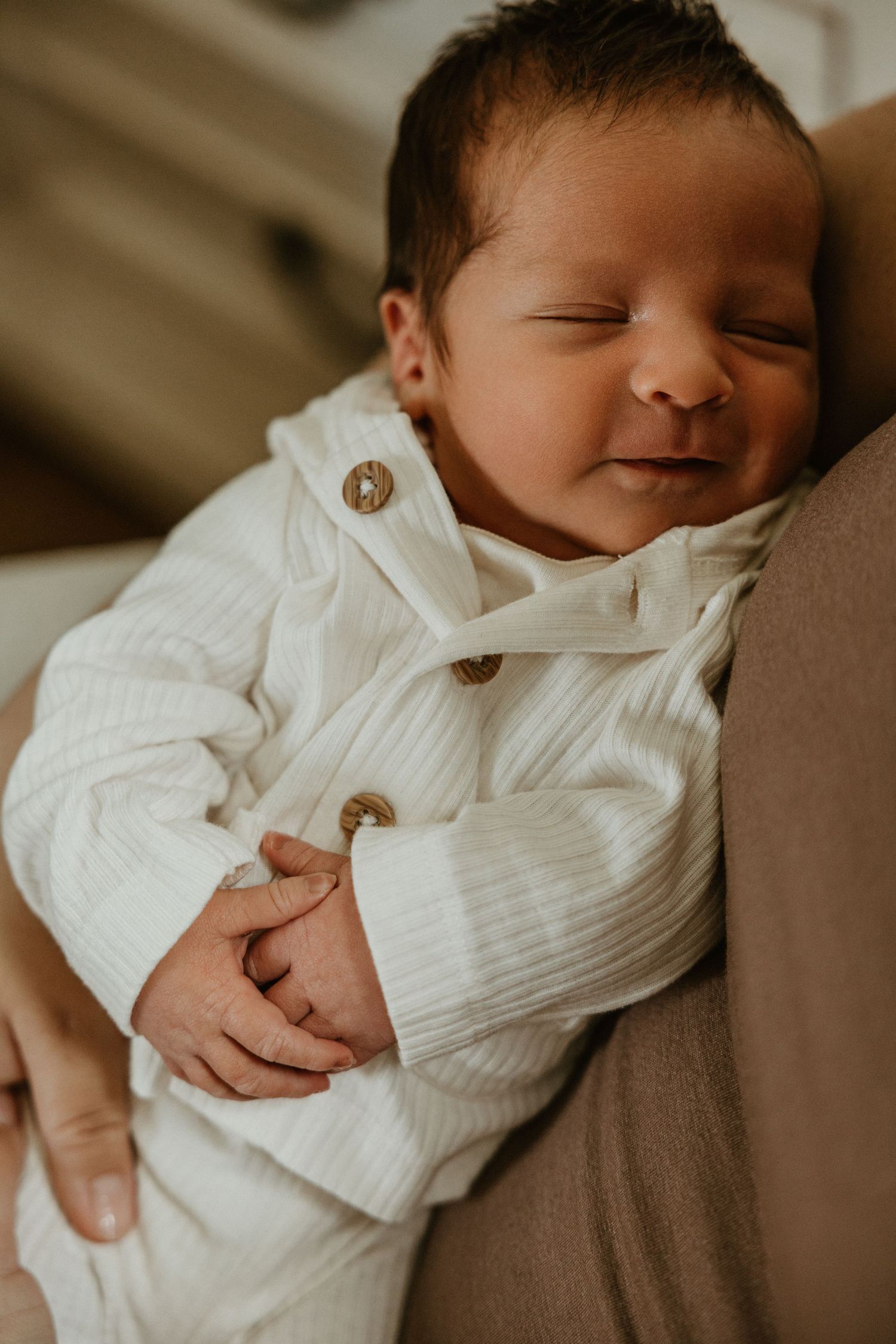 Newborn baby smiling in white ribbed button-up outfit, peacefully resting in someone's arms.