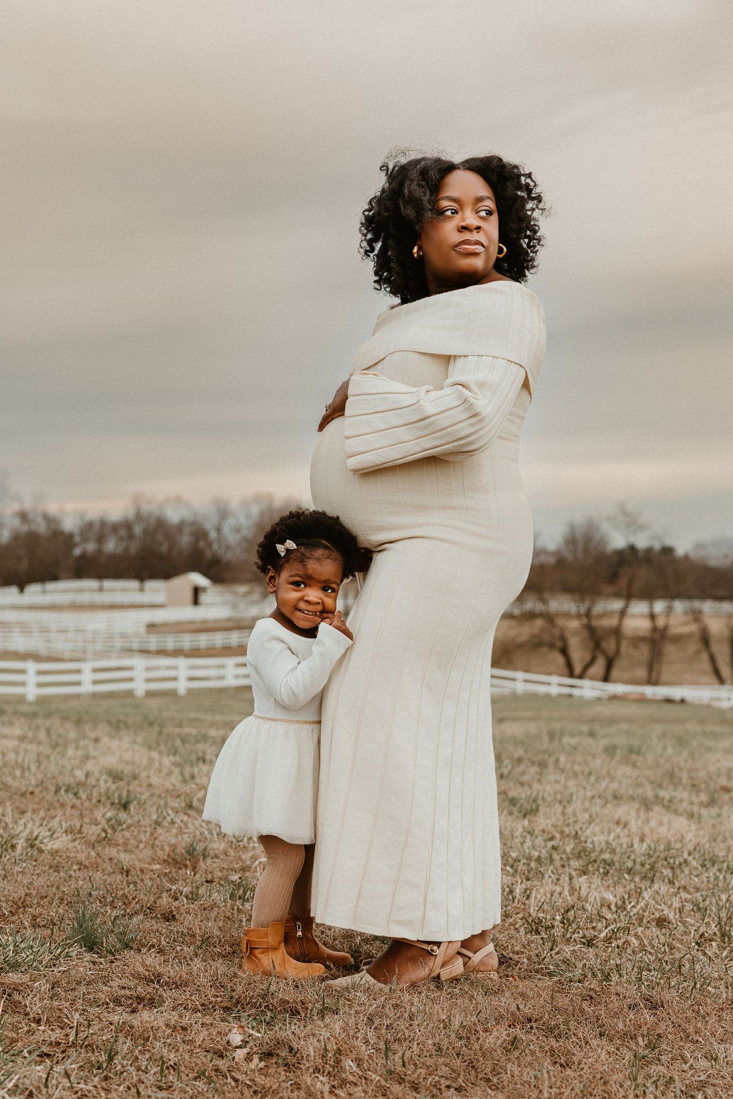 Pregnant Black woman in cream knit dress hugging toddler daughter outdoors at farm during maternity photoshoot.