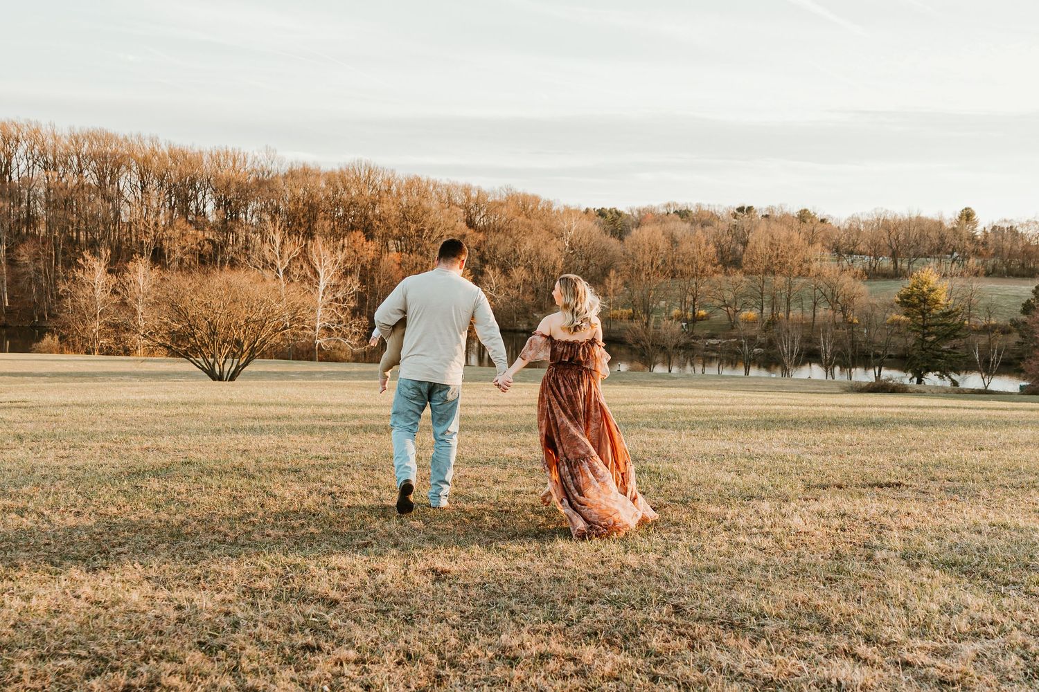 Couple holding hands walking through an open field with autumn trees in the background.