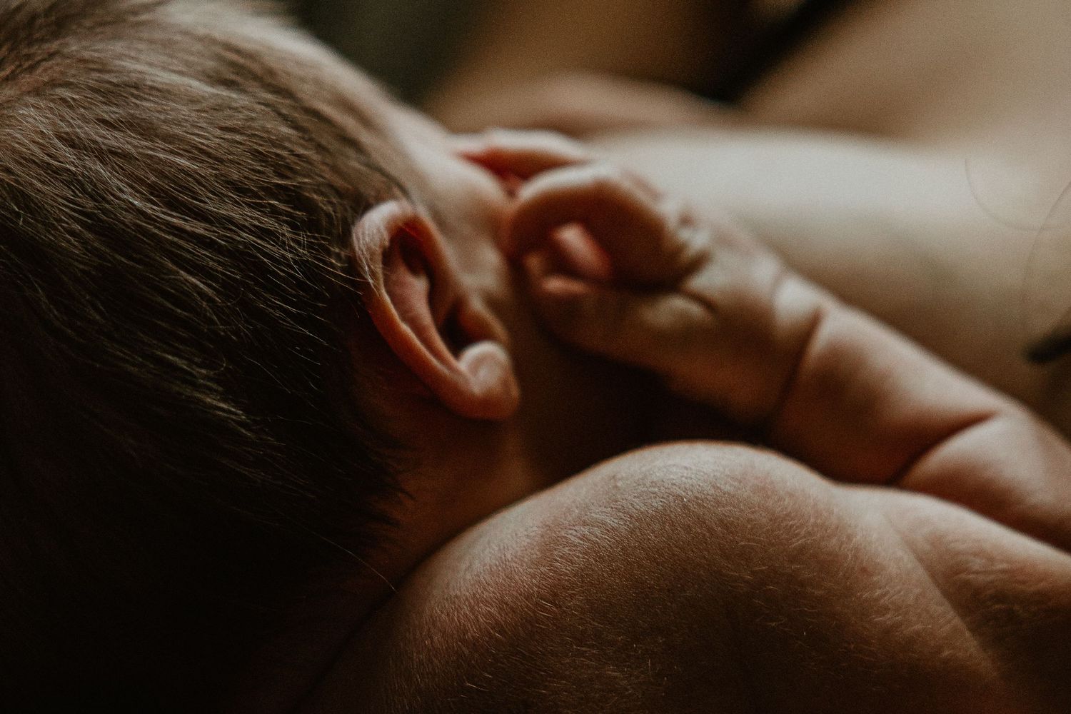 Close-up of a newborn baby's ear and tiny hand resting against skin in soft, warm light.