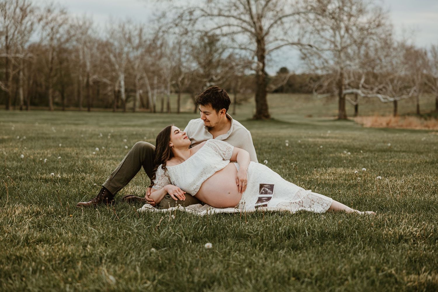 Pregnant couple sitting on grass in a park, sharing a tender moment with an ultrasound photo during a maternity photoshoot.