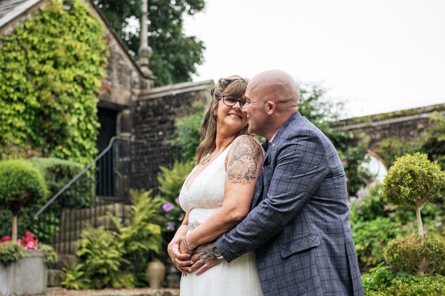 Bride and groom share a romantic embrace outdoors at a garden wedding venue, groom in blue suit kissing brides cheek.