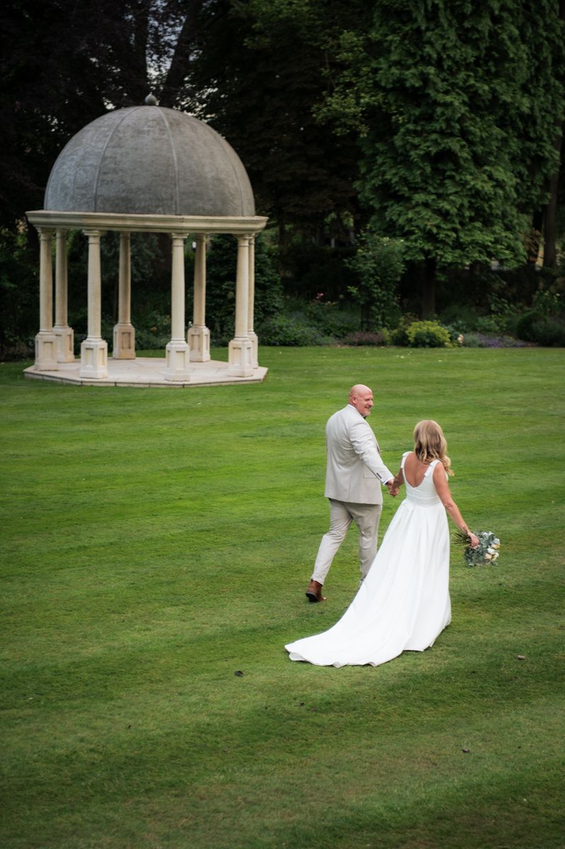 Bride and groom walk hand-in-hand across lush green lawn toward elegant stone gazebo in a beautiful garden setting.