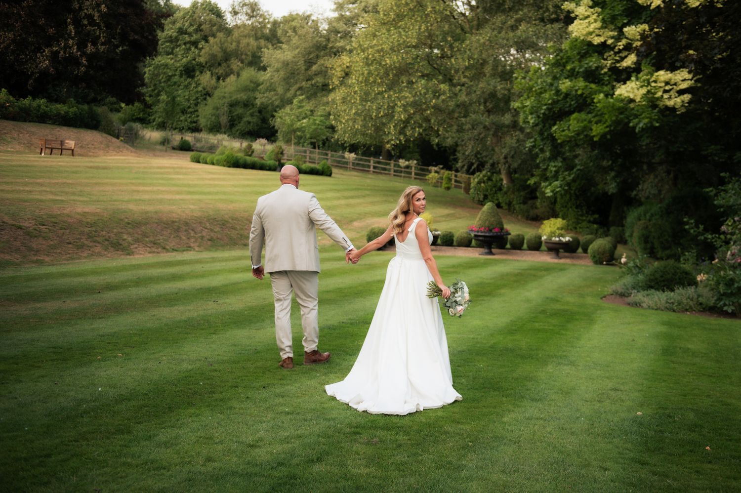 Bride and groom walking hand in hand across lush green lawn at outdoor wedding venue surrounded by trees.
